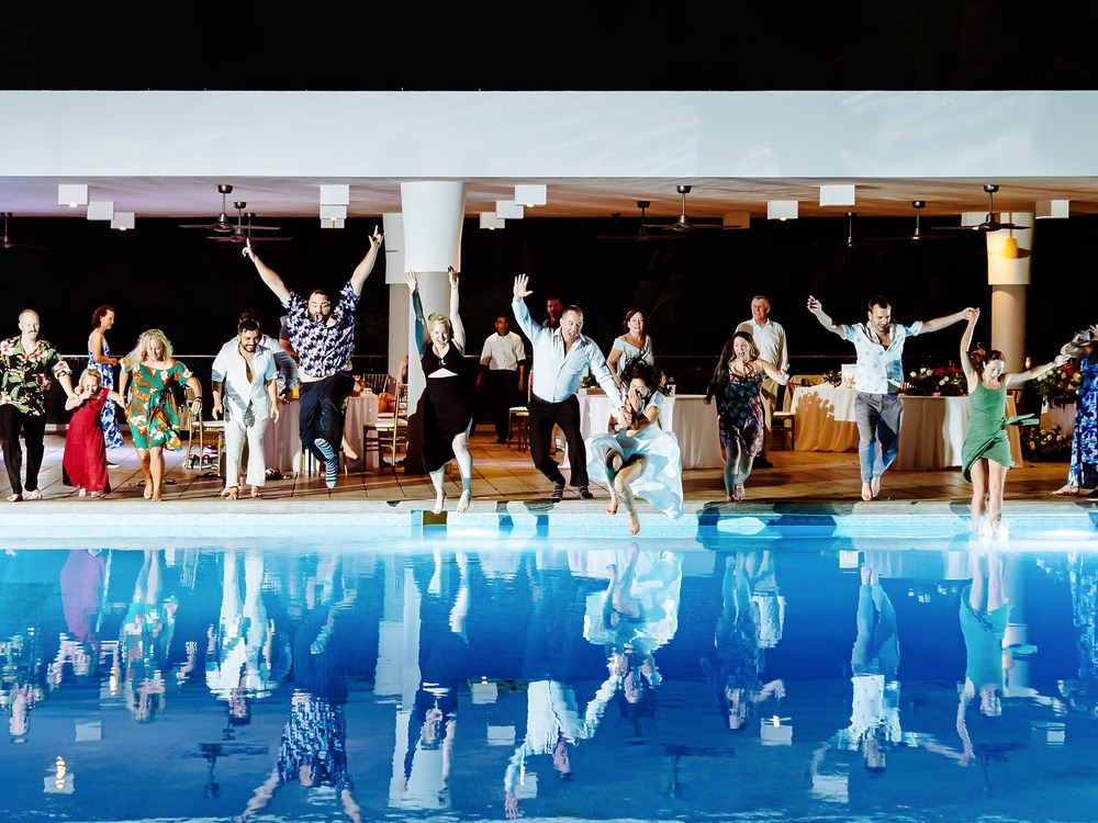 At a destination wedding celebration, a group of guests jumps into the swimming pool together in a burst of joyful excitement.