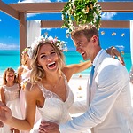 The smiling bride and groom share a joyful dance at their destination wedding, with the turquoise ocean creating a stunning beachside backdrop.