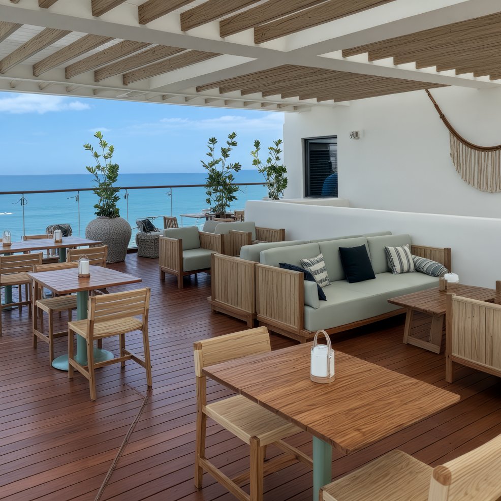 Terrace set for a destination wedding, with wooden tables, cushioned seats, ocean view, and pergola.