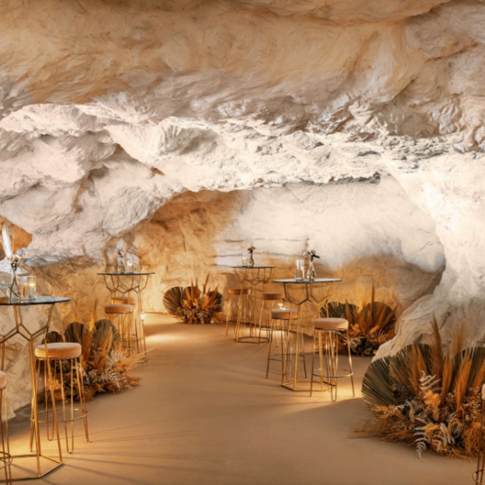 A cave-like room set for a destination wedding, featuring tall tables, stools, and dried floral decor.