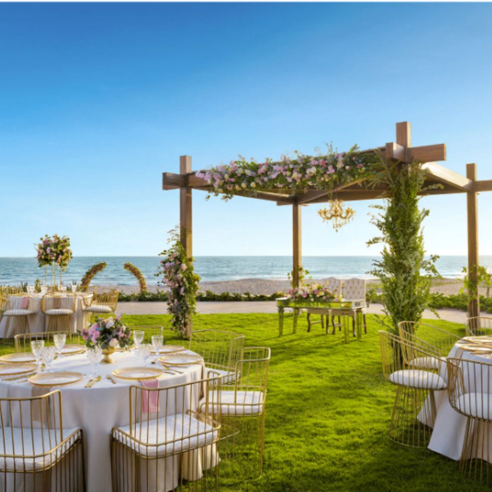 Destination wedding by the sea featuring decorated tables and a floral pergola under clear sunny skies.
