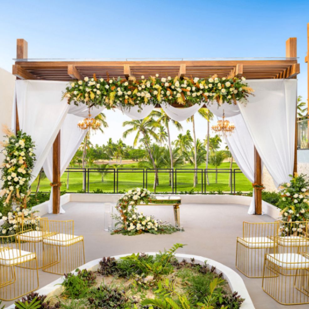 Destination wedding setup featuring floral decor, white drapes, and gold chairs beneath a wooden pergola.
