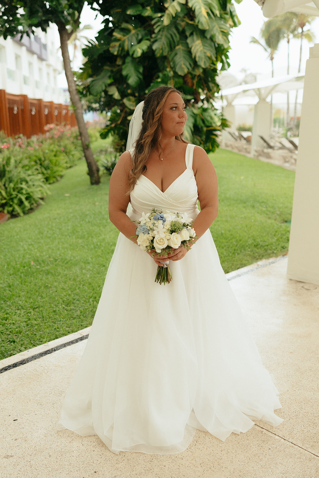 Bride in Stephanie and Mark (Copy) dress at destination wedding, holding bouquet near greenery, buildings.