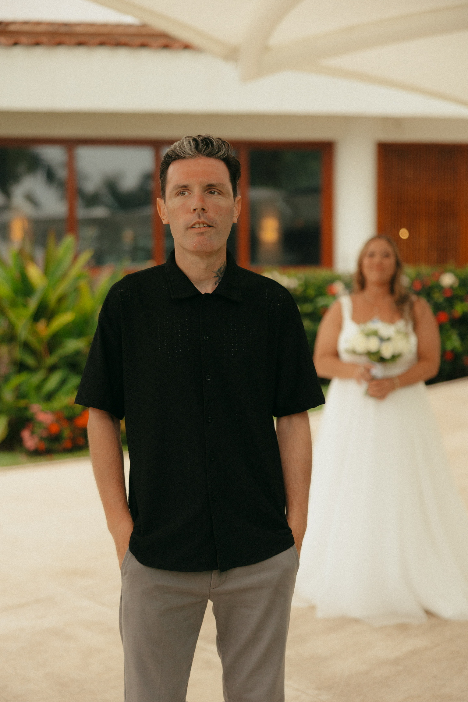 At their destination wedding, a man stands in front with a woman in a wedding dress behind him.