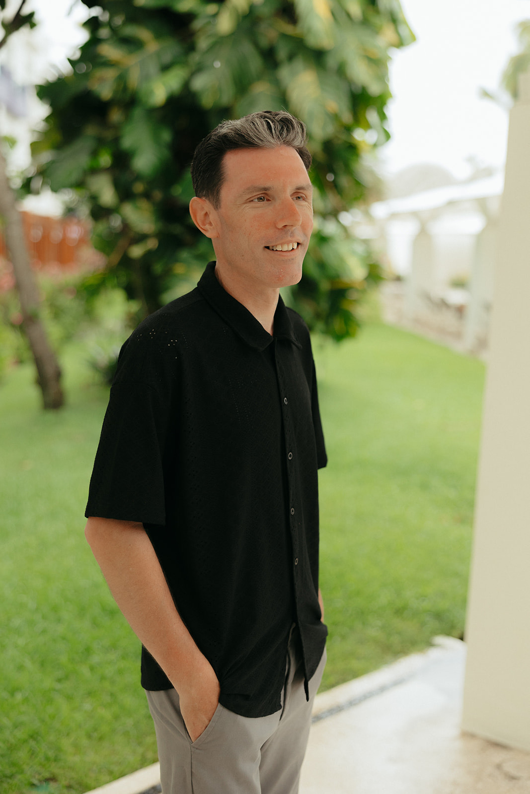Man in "Stephanie and Mark (Copy)" shirt outside, hands in pockets, with lush greenery—wedding setting.
