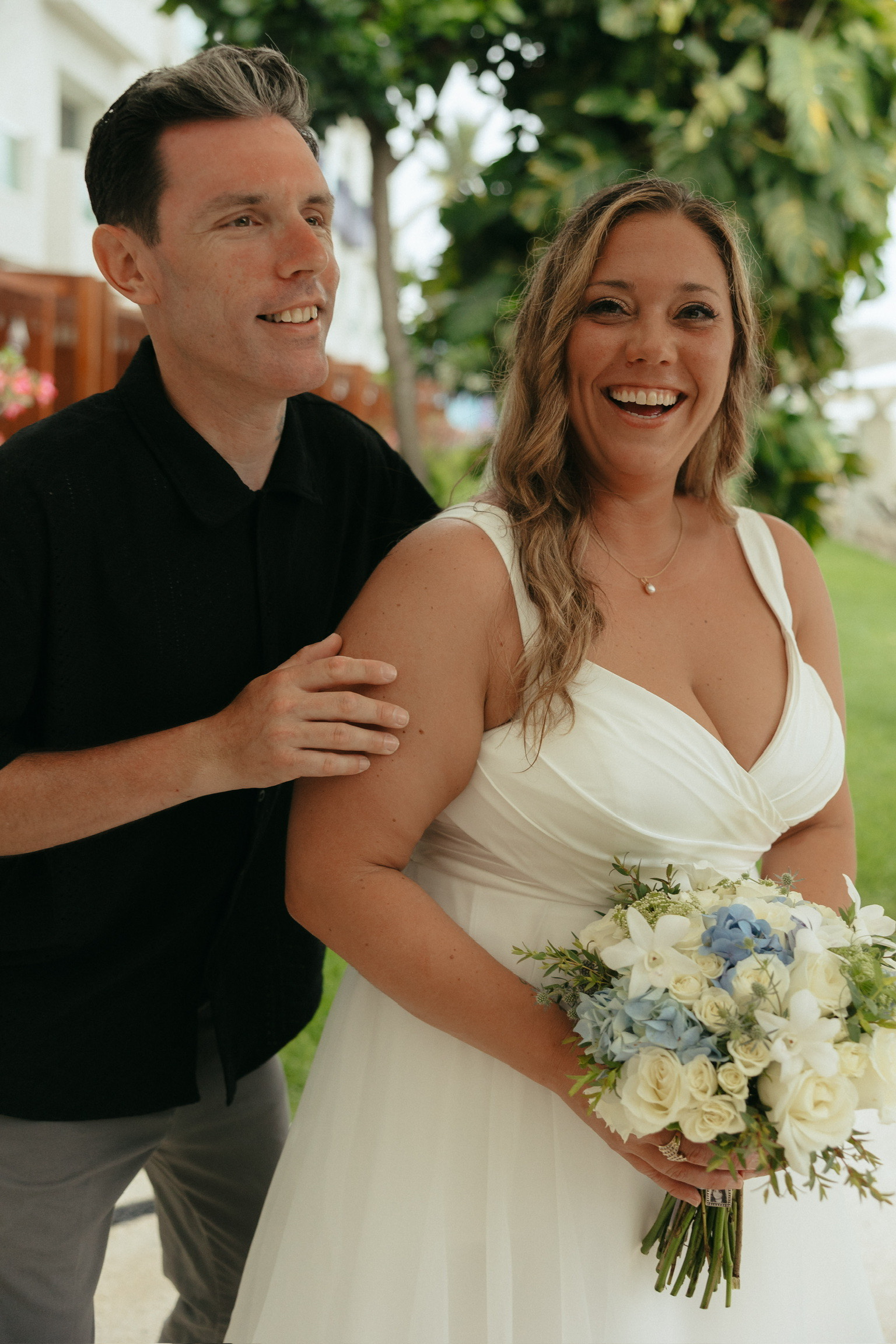 Stephanie and Mark smile outdoors at their destination wedding; she wears white and carries a bouquet.