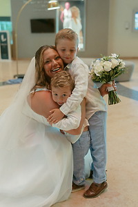 At a destination wedding, the bride in white hugs two boys, one of whom is holding white flowers.