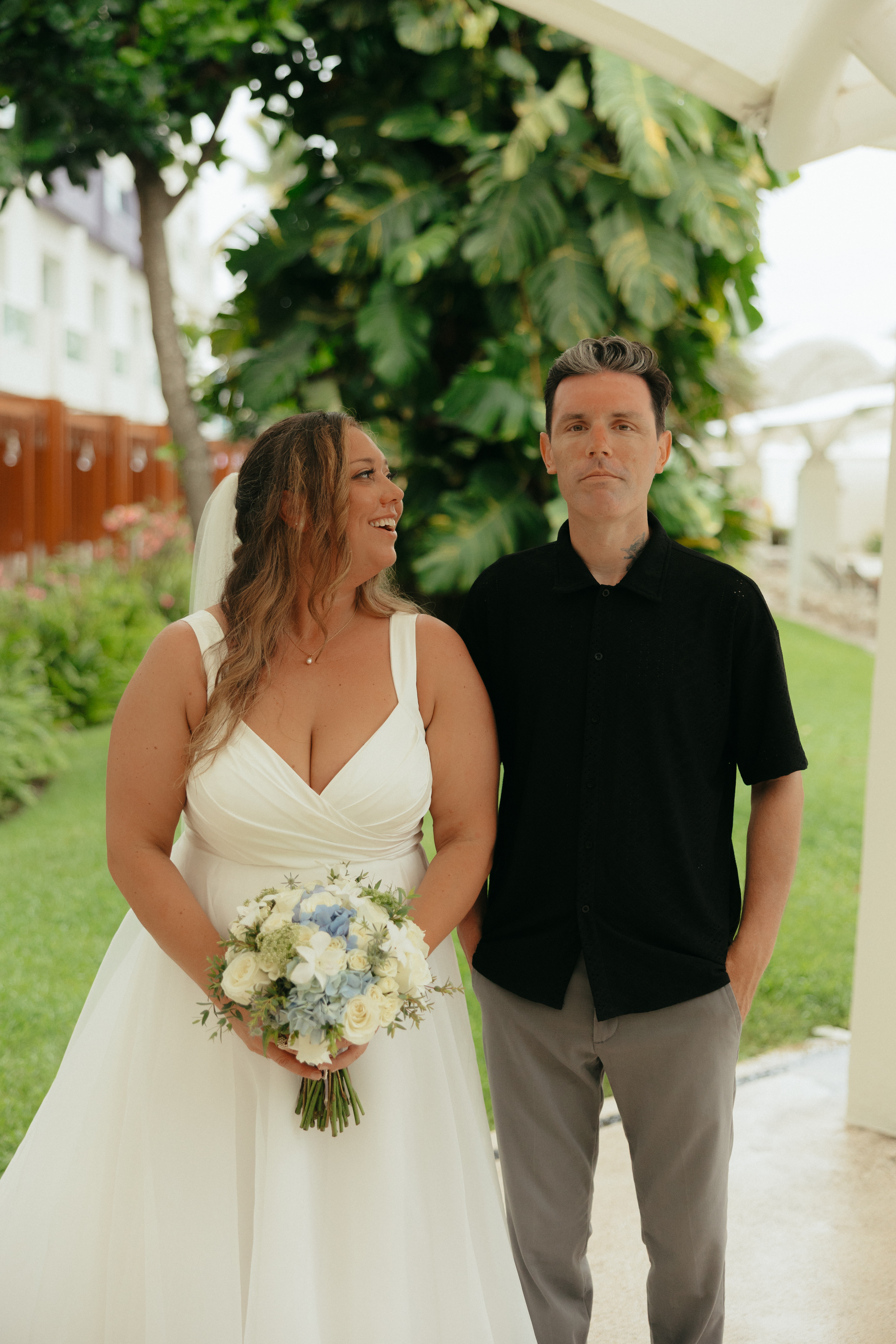 Bride in white dress holding bouquet stands beside man in black shirt at outdoor destination wedding.