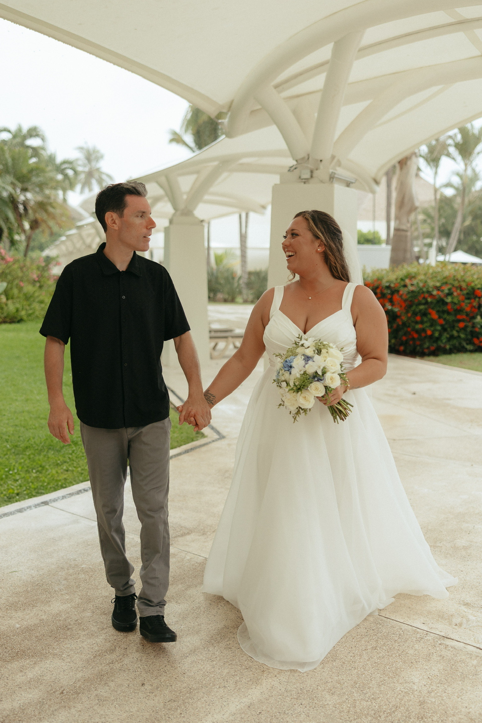 Stephanie and Mark hold hands outdoors at a destination wedding, smiling—she in a white dress, he casual.