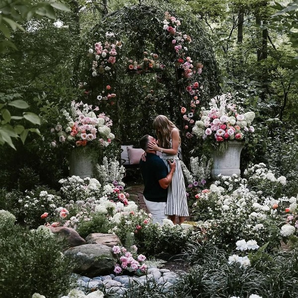 A man proposes on one knee to a woman in a lush, flower-filled garden, perfect for a destination wedding.