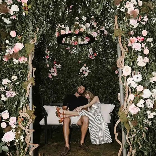 A couple sits on a bench under a wedding floral archway decorated with greenery and hanging flowers.