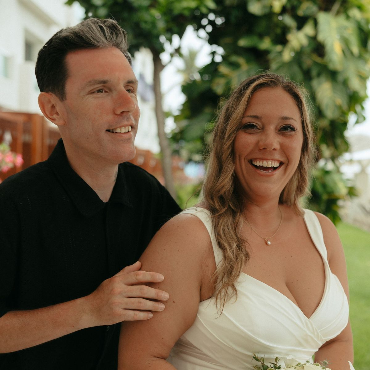 Stephanie and Mark smile at the camera outdoors at their destination wedding; Stephanie holds flowers in her white dress.