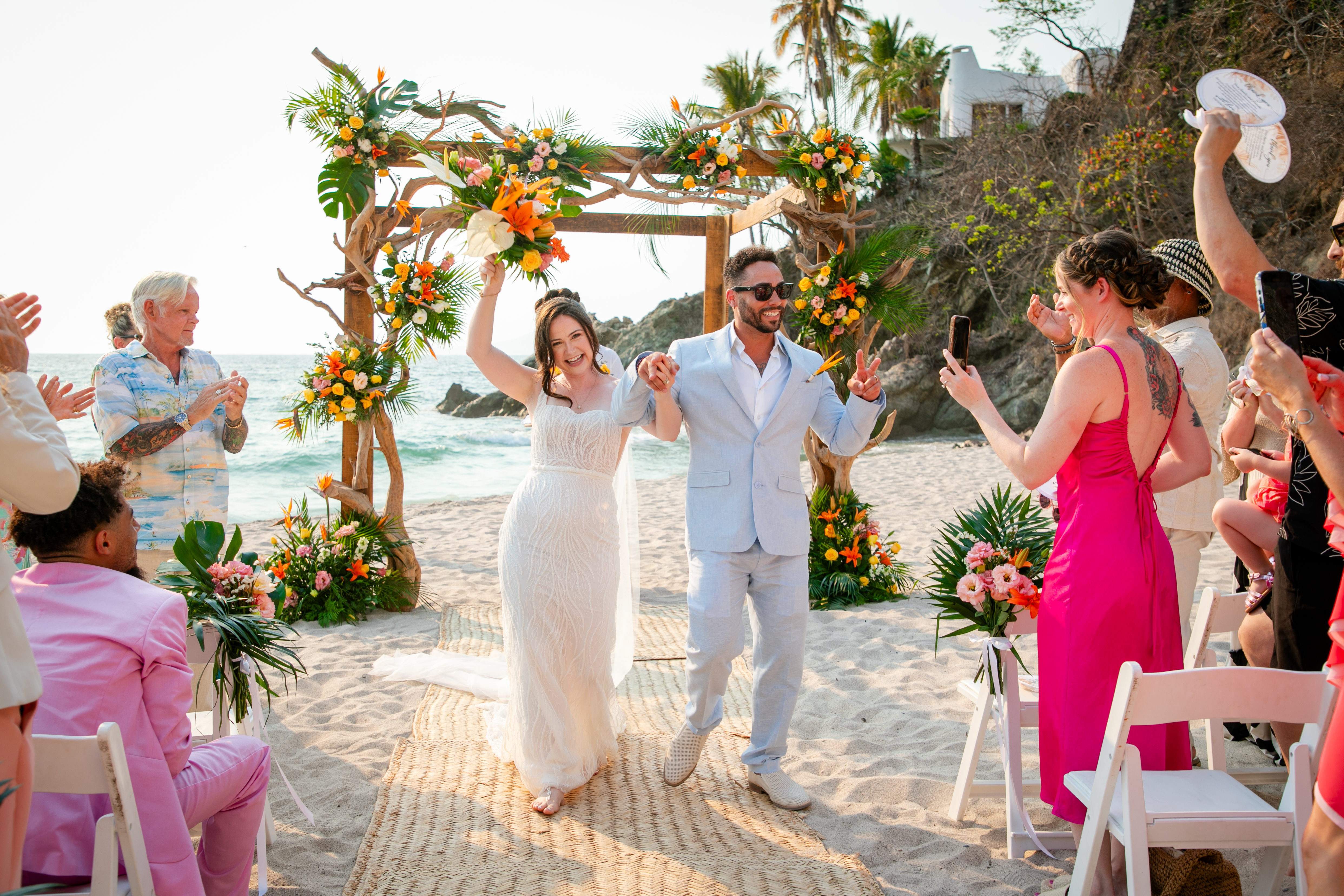 Jordan and Kievan stroll hand-in-hand down the sandy aisle after their destination wedding, surrounded by cheering guests.