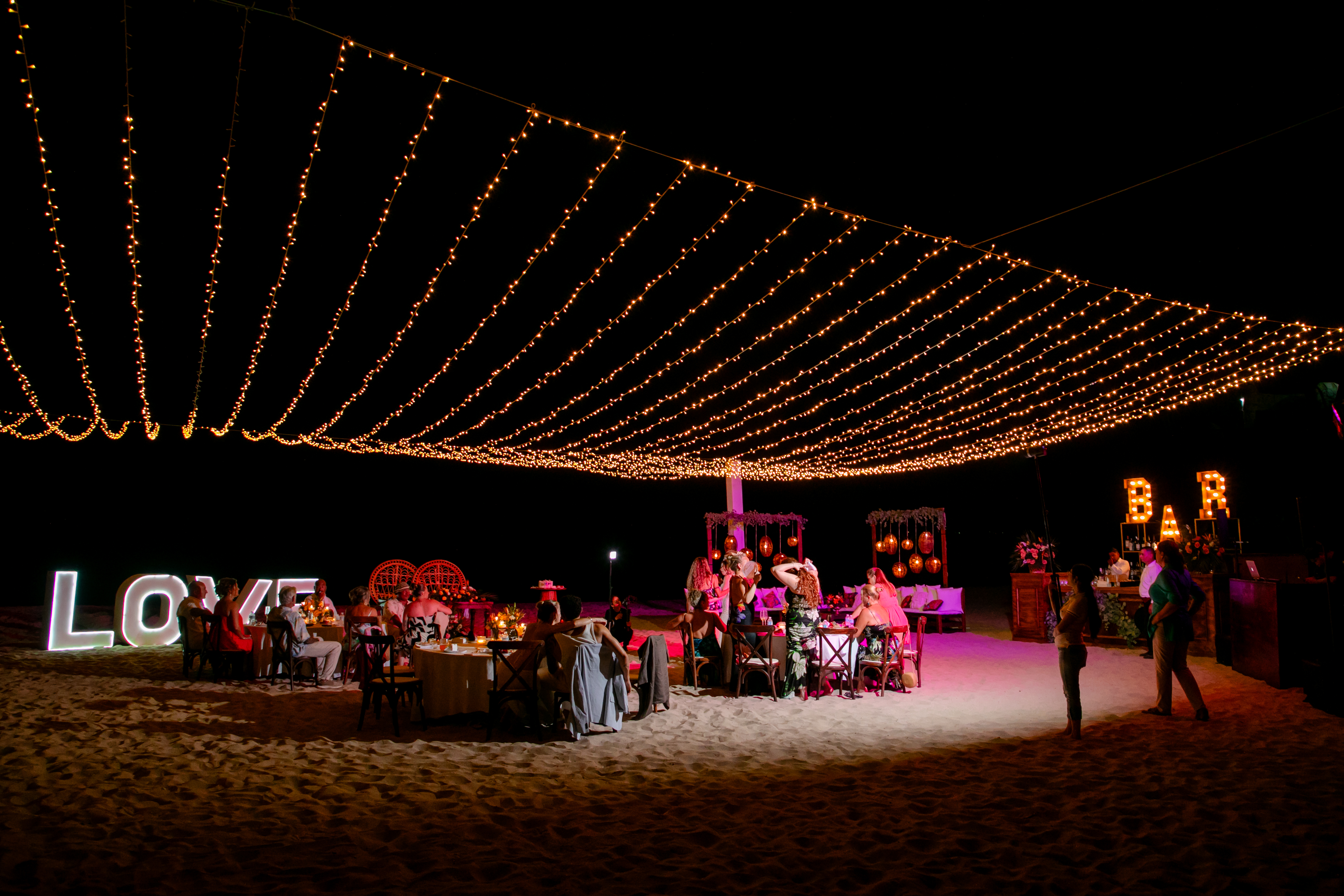 Nighttime destination wedding on the beach, with guests gathered under twinkling string lights and a large "LOVE" sign, celebrating Jordan and Kievan.