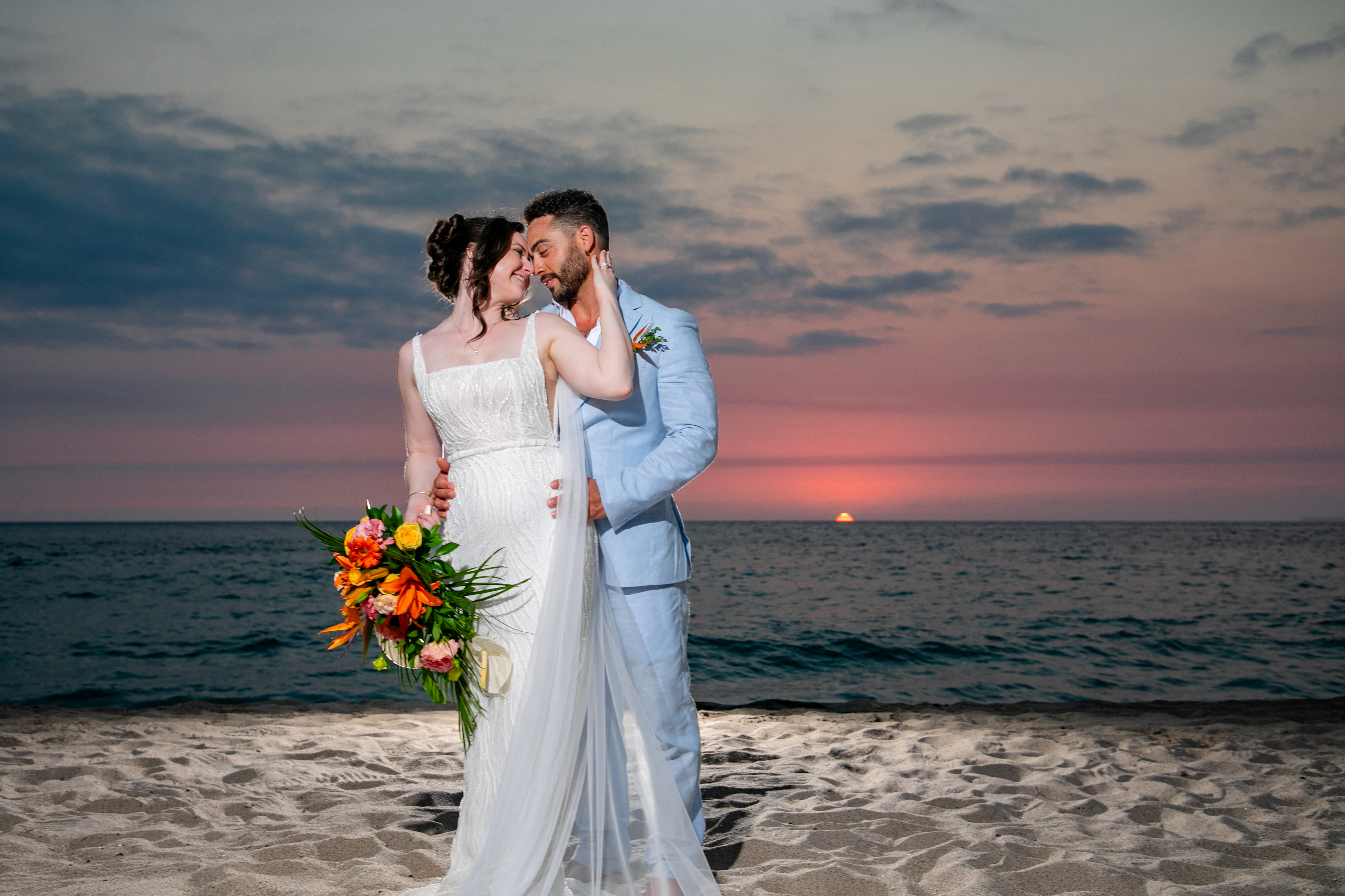 Jordan and Kievan embrace on the beach at sunset during their destination wedding, holding a vibrant bouquet as waves gently lap the shore.