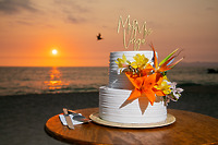 Jordan and Kievan’s romantic destination wedding featured a two-tier white wedding cake adorned with vibrant orange flowers, beautifully set by the beach at sunset.