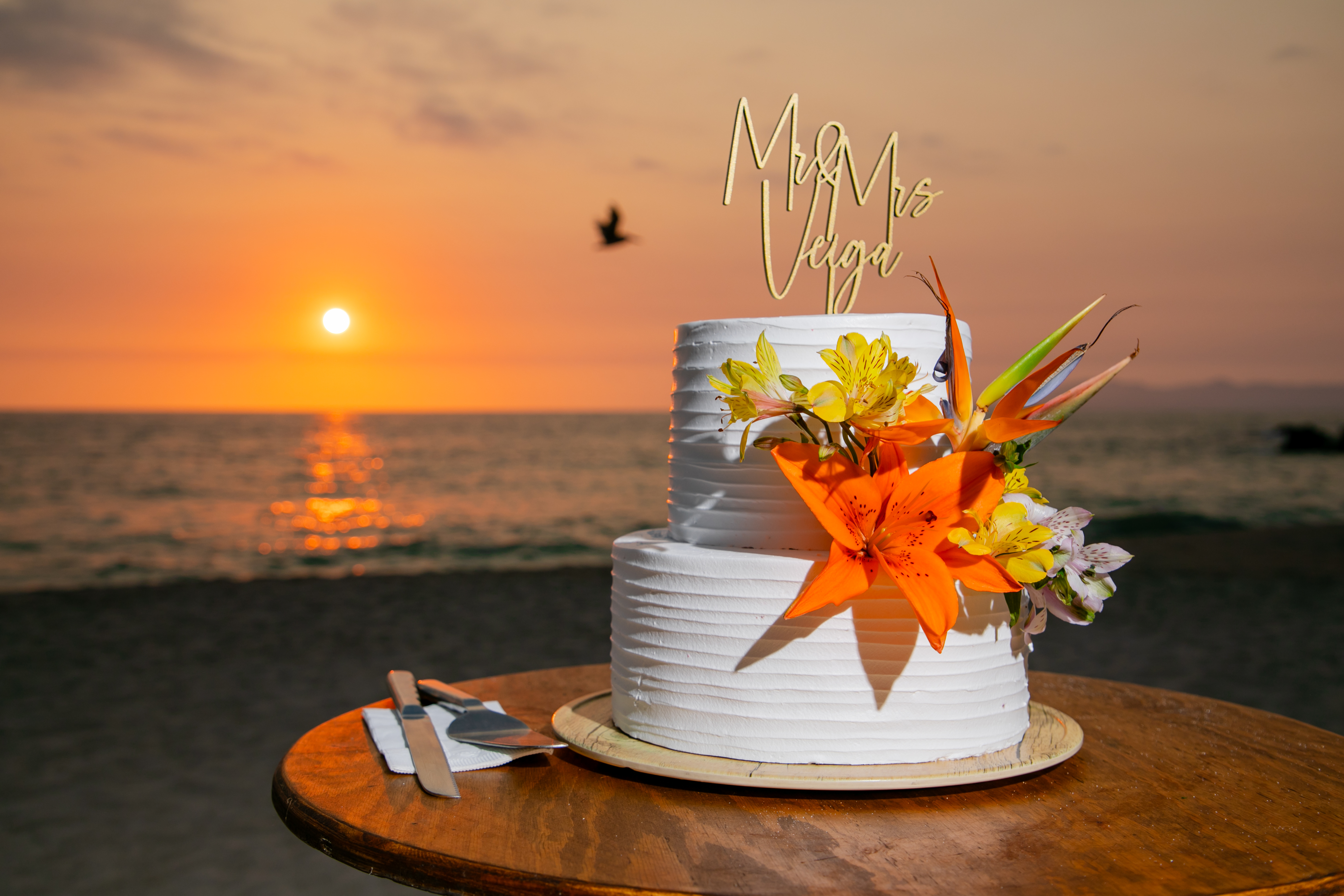 Jordan and Kievan’s romantic destination wedding featured a two-tier white wedding cake adorned with vibrant orange flowers, beautifully set by the beach at sunset.