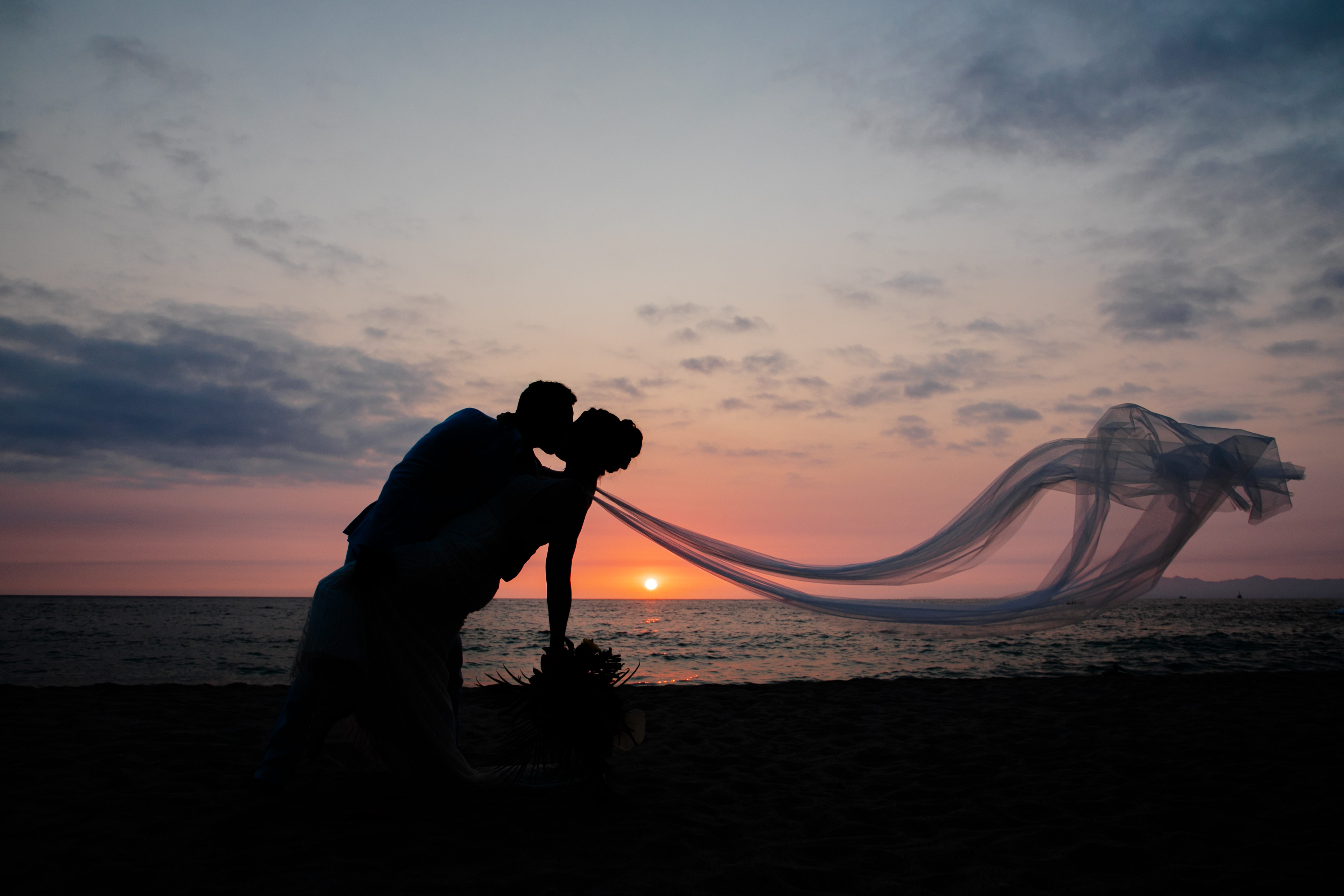 Jordan and Kievan (Copy): A couple shares a romantic kiss on the beach at sunset during their destination wedding, the bride's veil flowing gracefully in the wind.