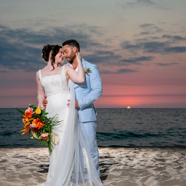 Jordan and Kievan embrace on a beach at sunset during their destination wedding, holding a vibrant bouquet of flowers.