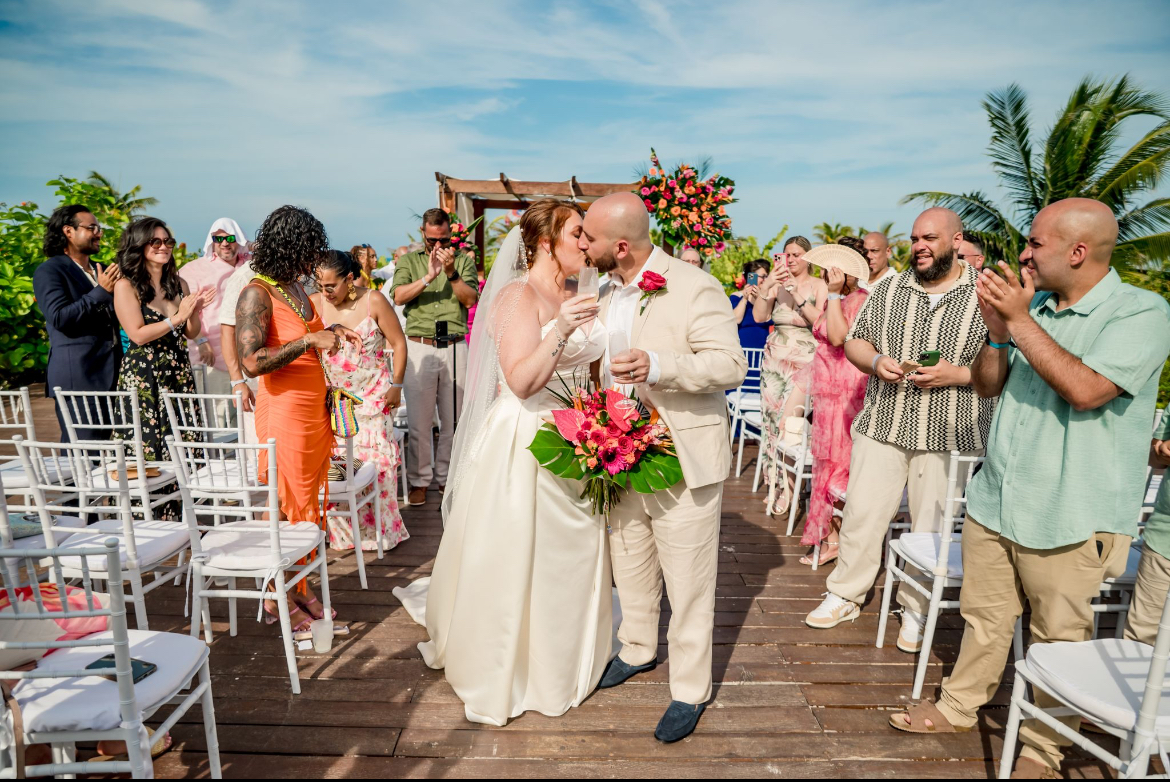 At their destination wedding, Mobolaji and Sylvia kiss as guests applaud on a bright, sunny day outdoors.