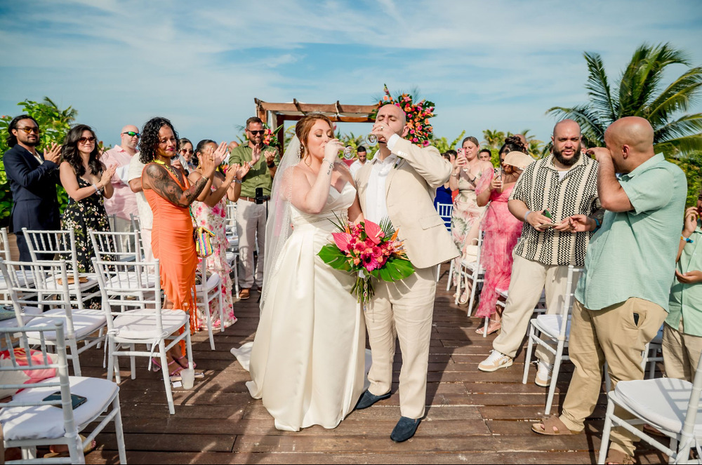Mobolaji and Sylvia sip drinks together as they walk down the aisle at their outdoor destination wedding.