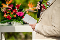 Beige-suited wedding host with microphone and vow booklet, vibrant flowers accent a destination wedding.