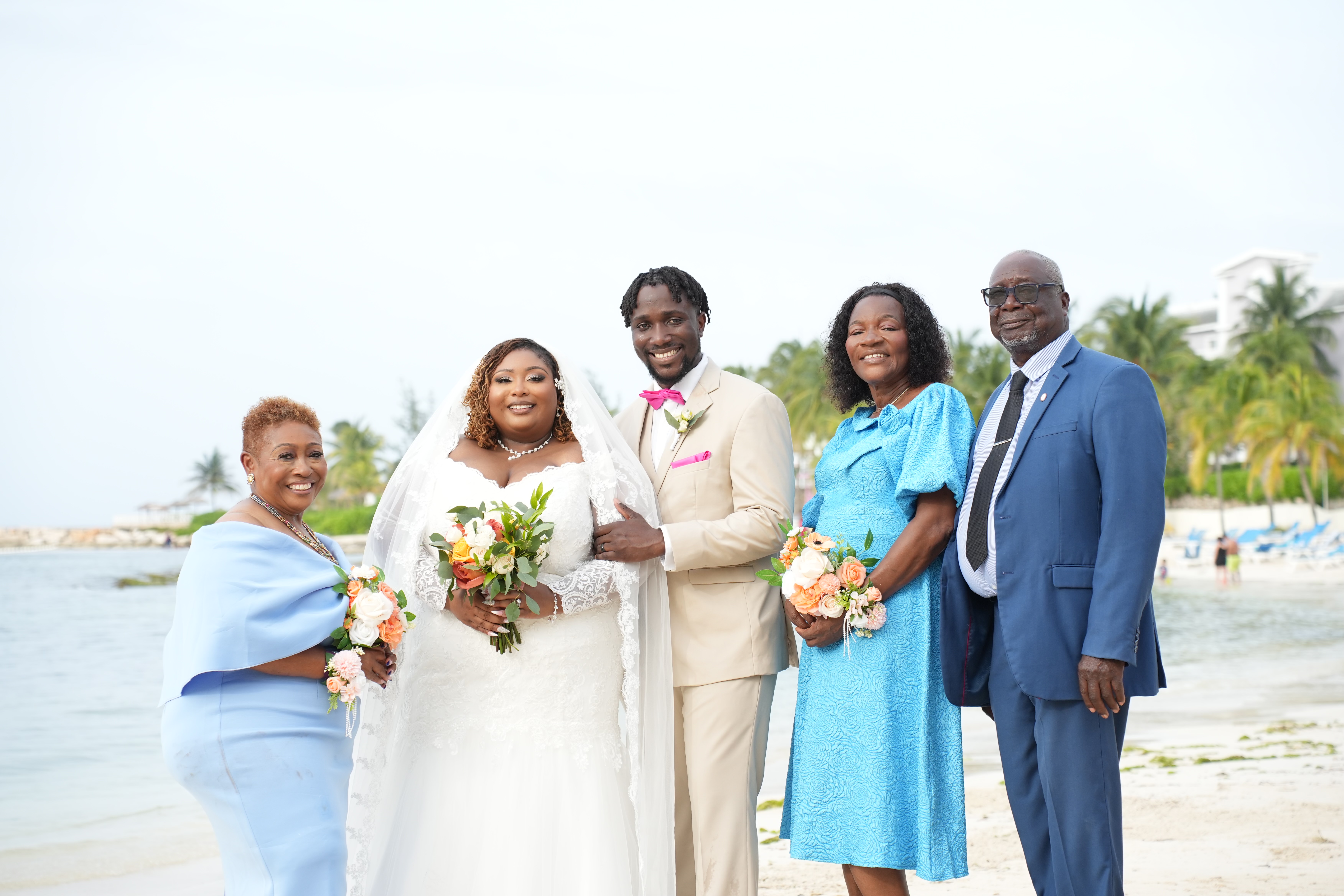 Mallory and Kharey pose with three guests on the beach, all elegantly dressed in formal attire for their destination wedding.