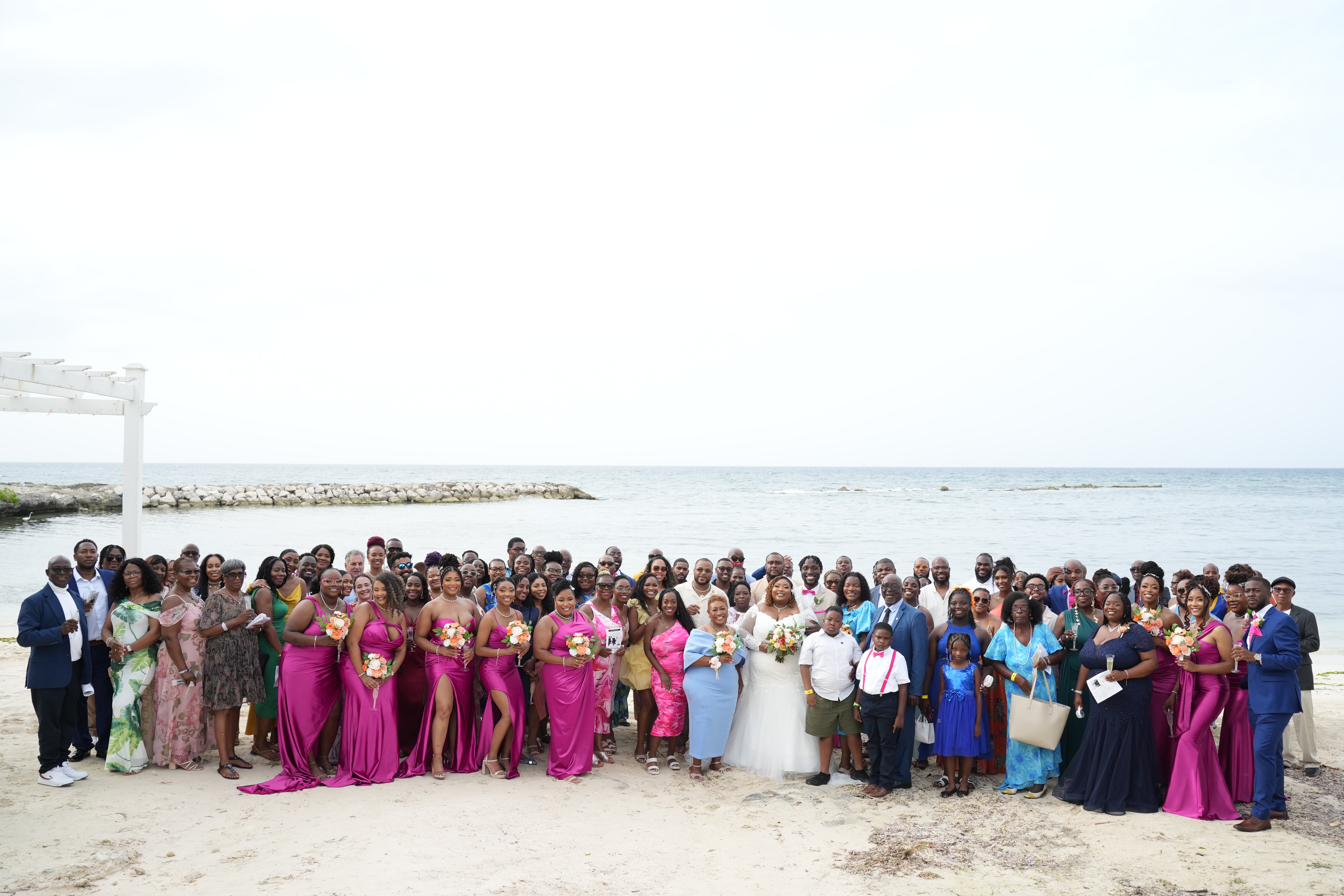 Mallory and Kharey’s wedding party gathers on the sandy beach, with the ocean providing a breathtaking backdrop for their destination wedding celebration.