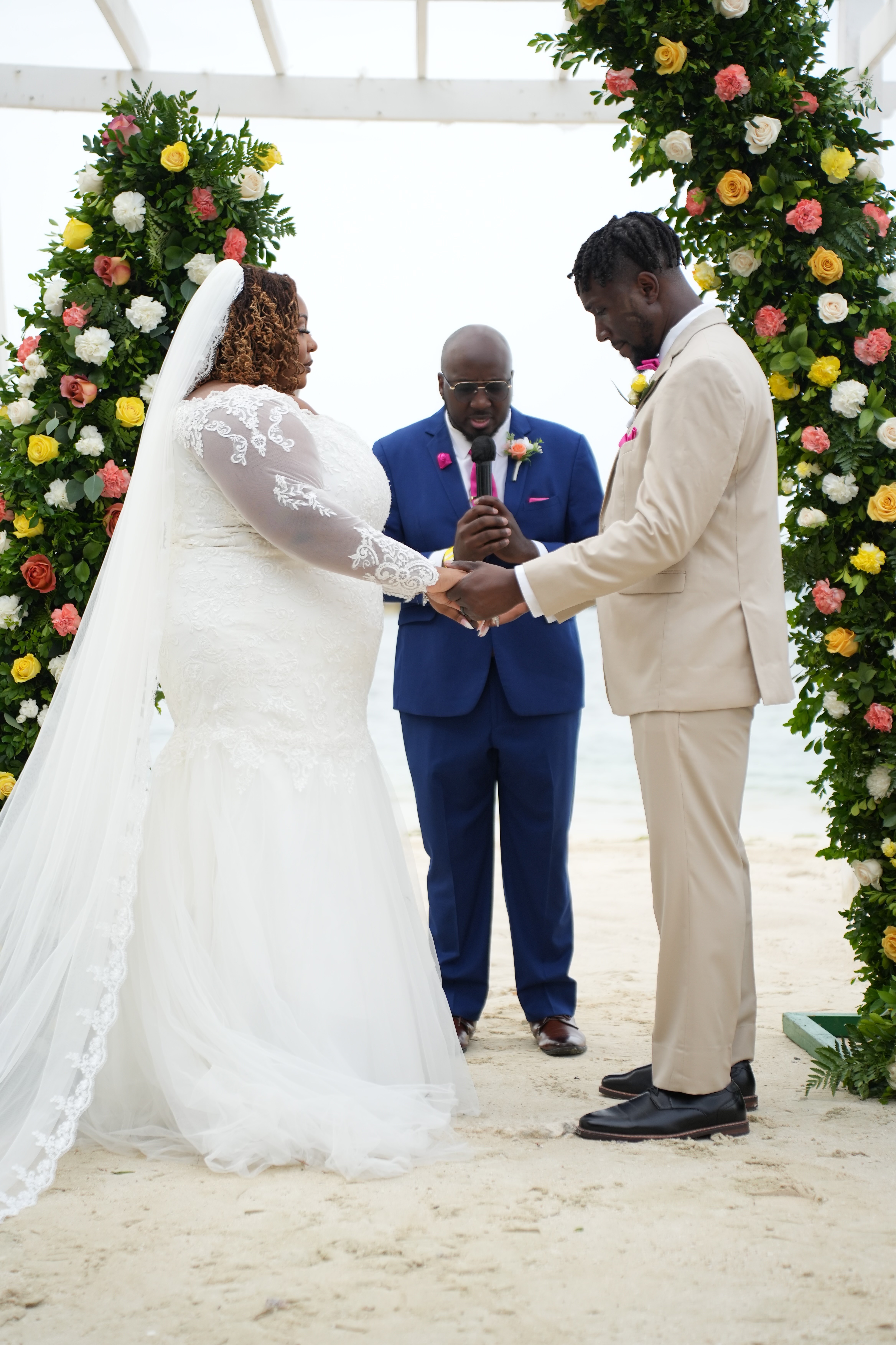 Kiah and Rodney hold hands during their destination wedding ceremony on the beach, surrounded by floral arches and joined by an officiant.