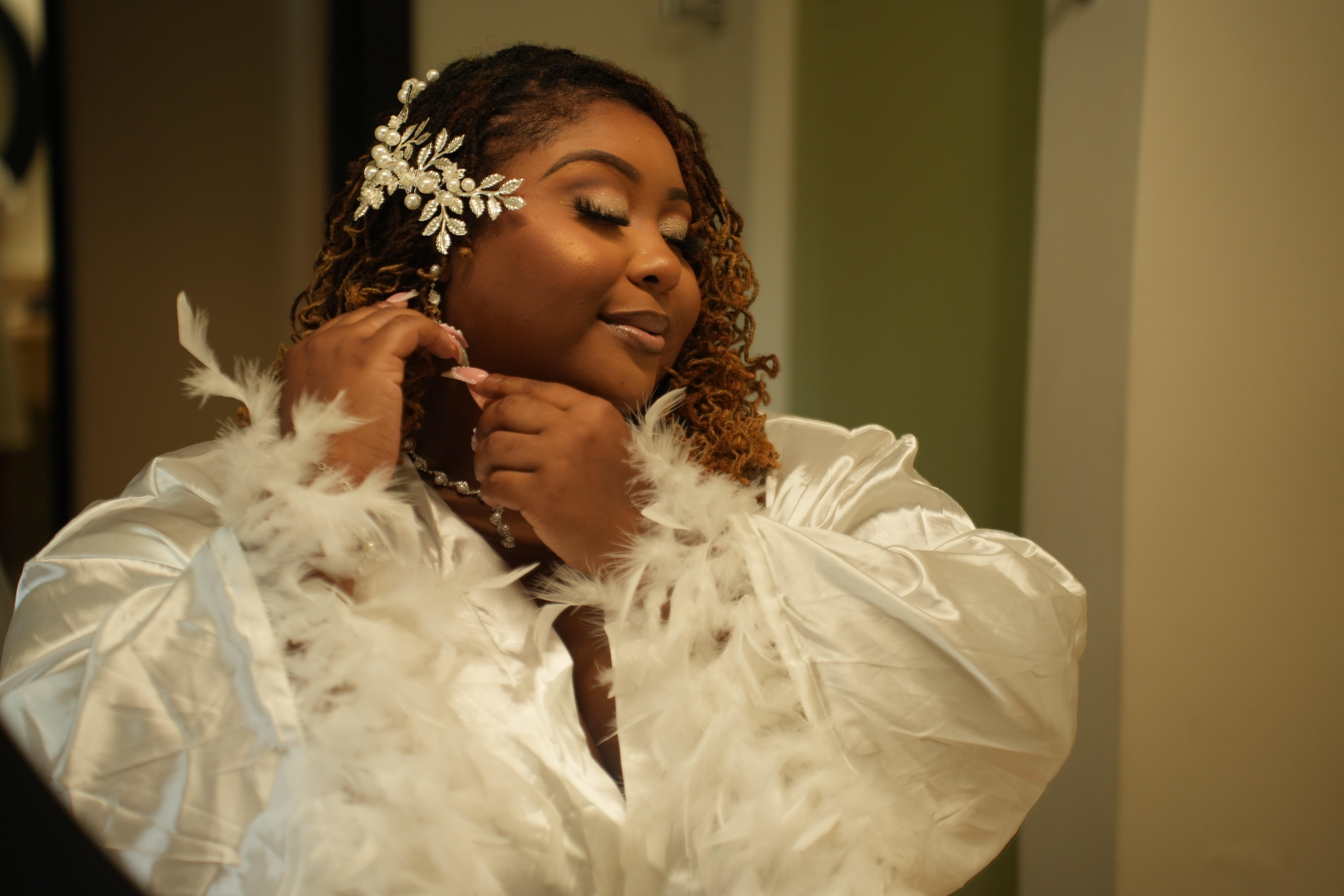 A woman in a white Kiah and Rodney robe, adorned with feather trim and an elegant hairpiece, adjusts her earring as she prepares for the destination wedding.