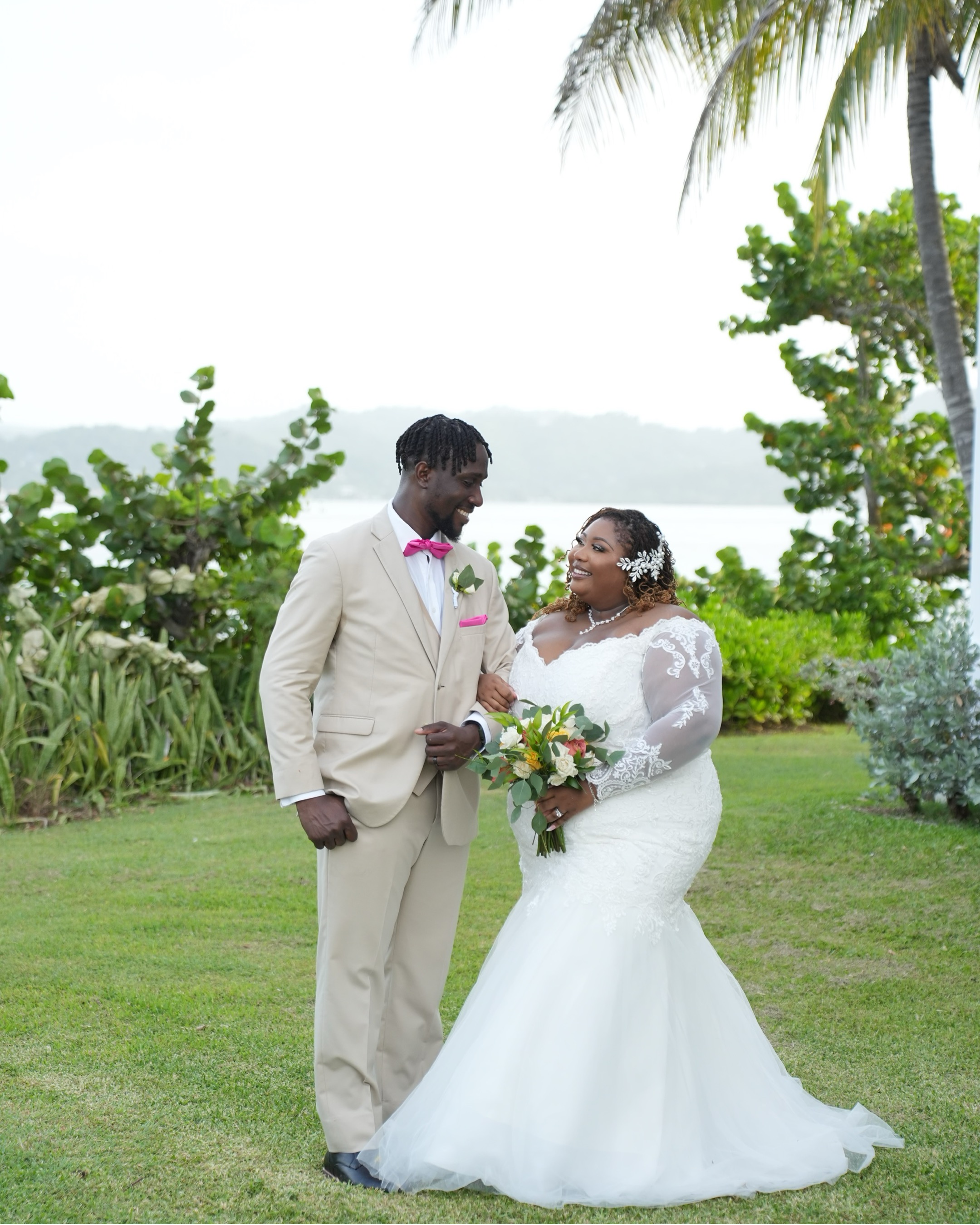 Kiah and Rodney smile together outdoors at their destination wedding, with lush greenery and shimmering water in the background.