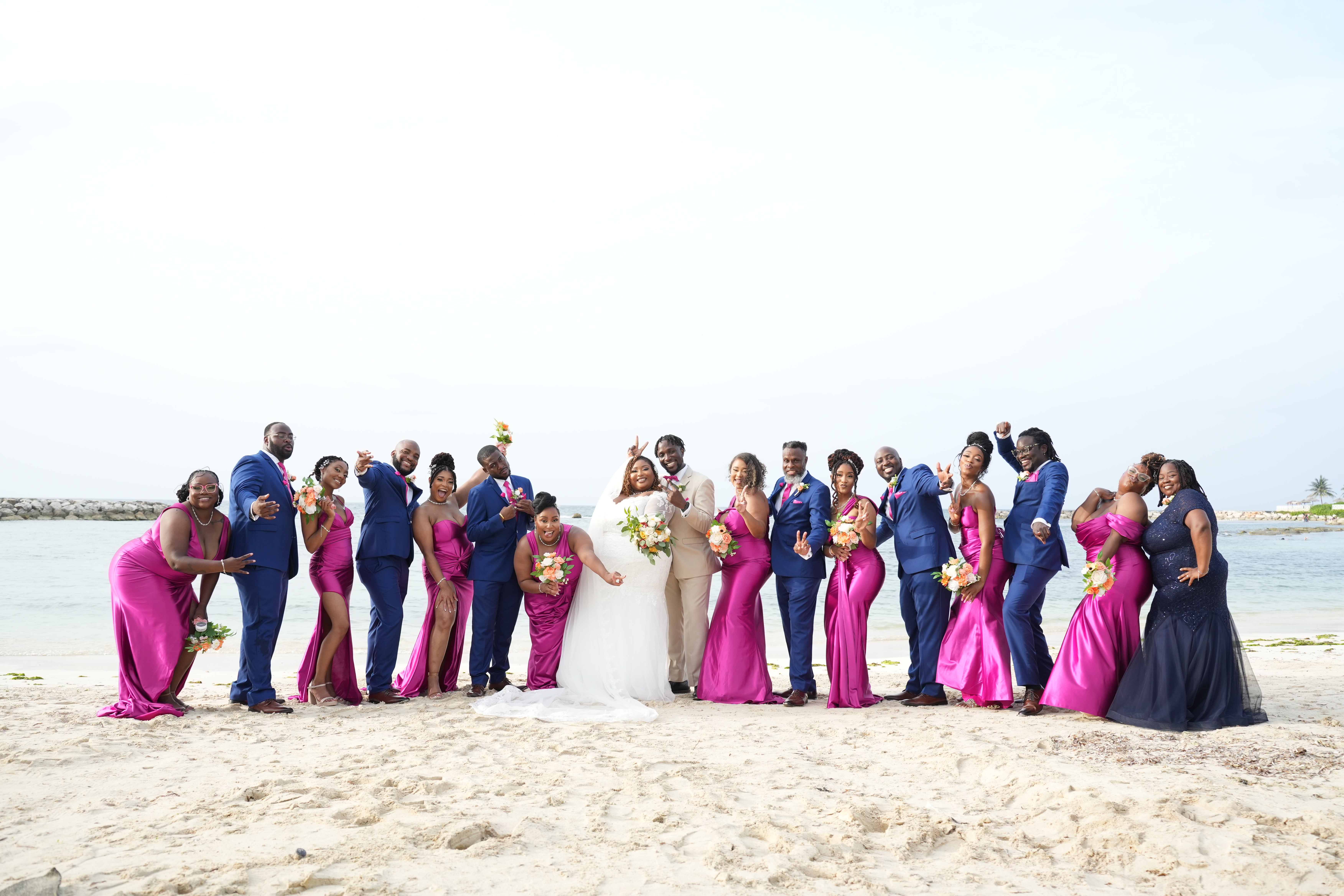 The wedding party for Kiah and Rodney poses on a sandy beach in vibrant magenta and blue outfits, beautifully capturing the spirit of their destination wedding.