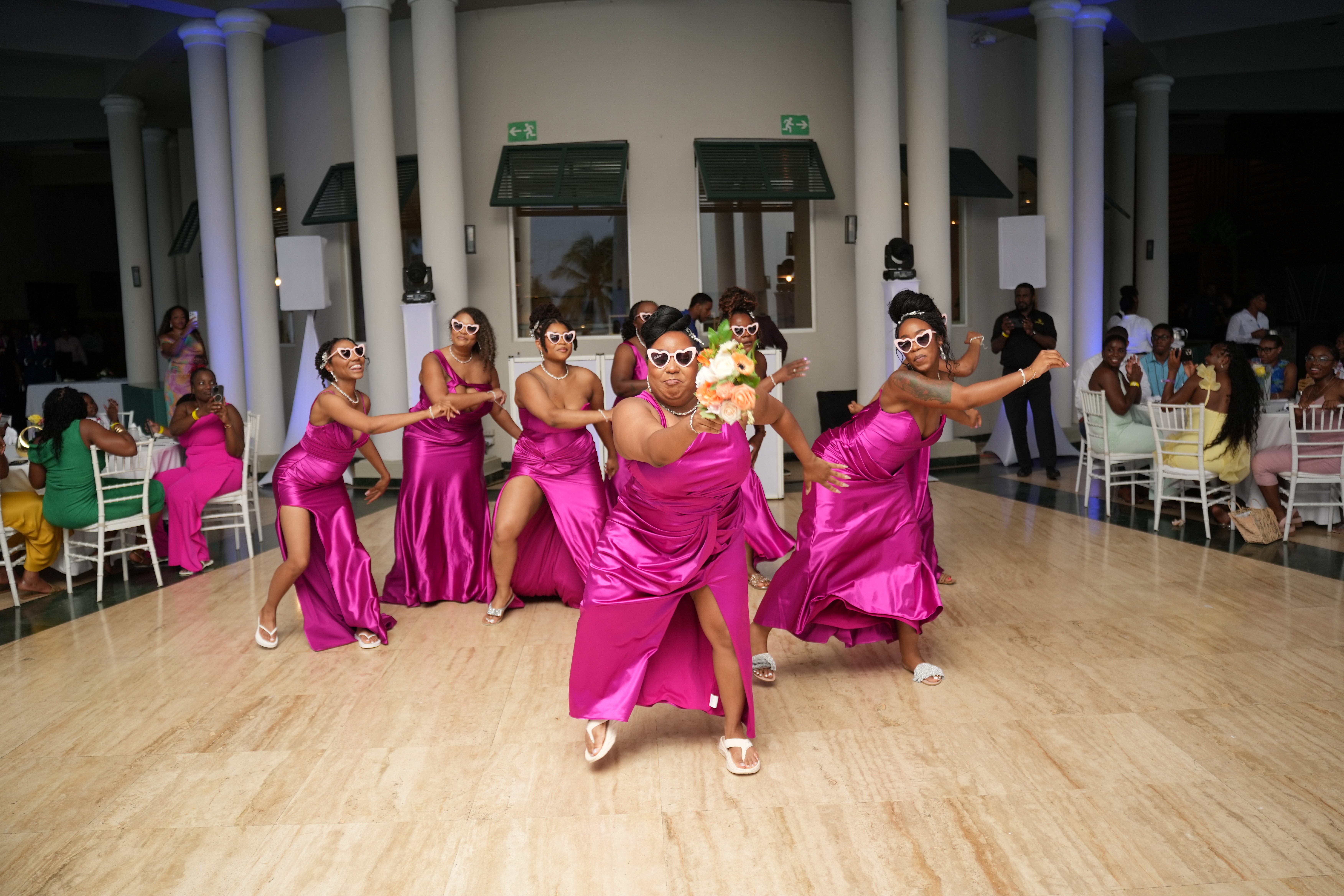 Six women in pink dresses and sunglasses dance at Kiah and Rodney’s destination wedding, entertaining seated guests.