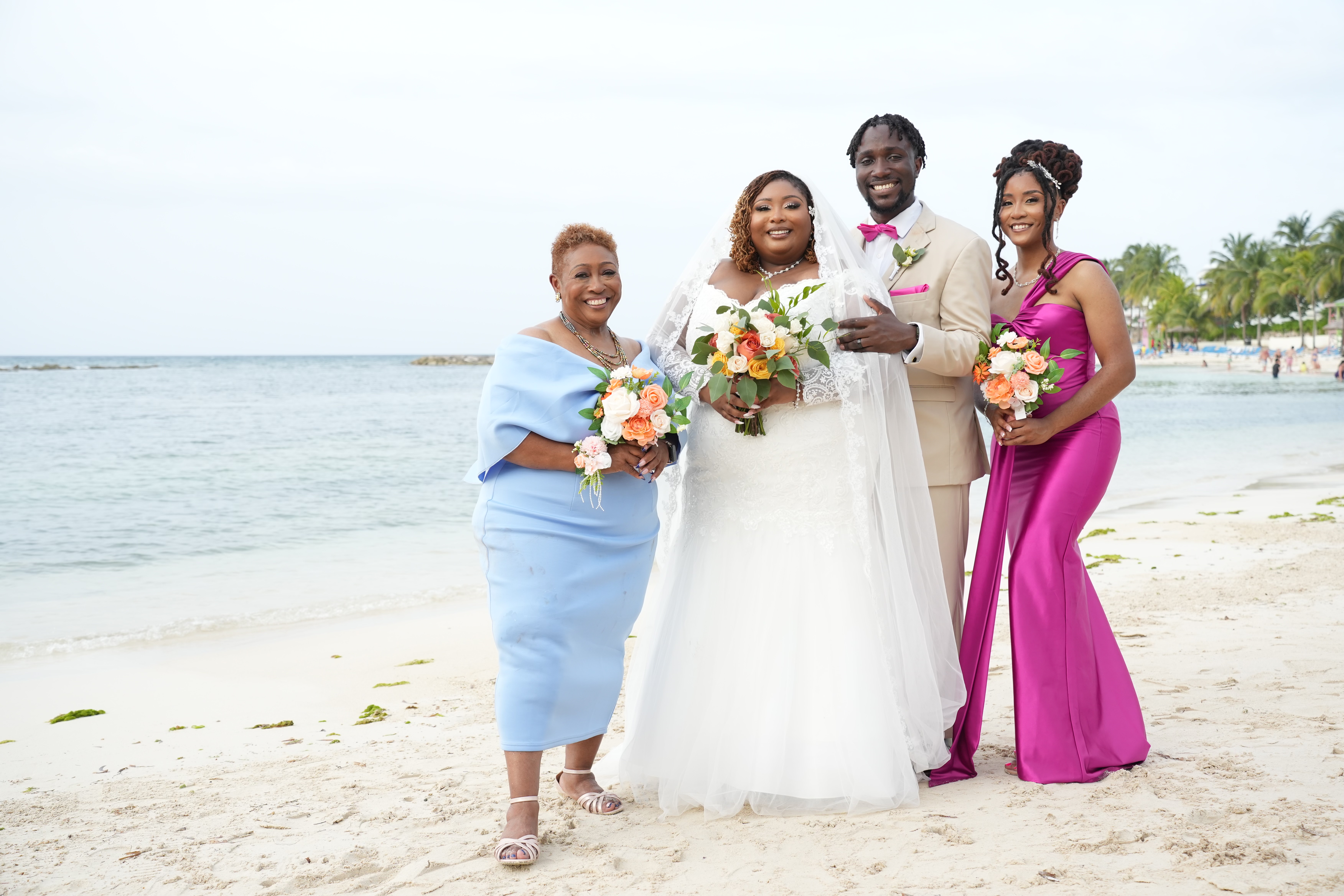 Four people dressed in elegant wedding attire smile and hold bouquets on a picturesque beach, celebrating Kiah and Rodney’s destination wedding.