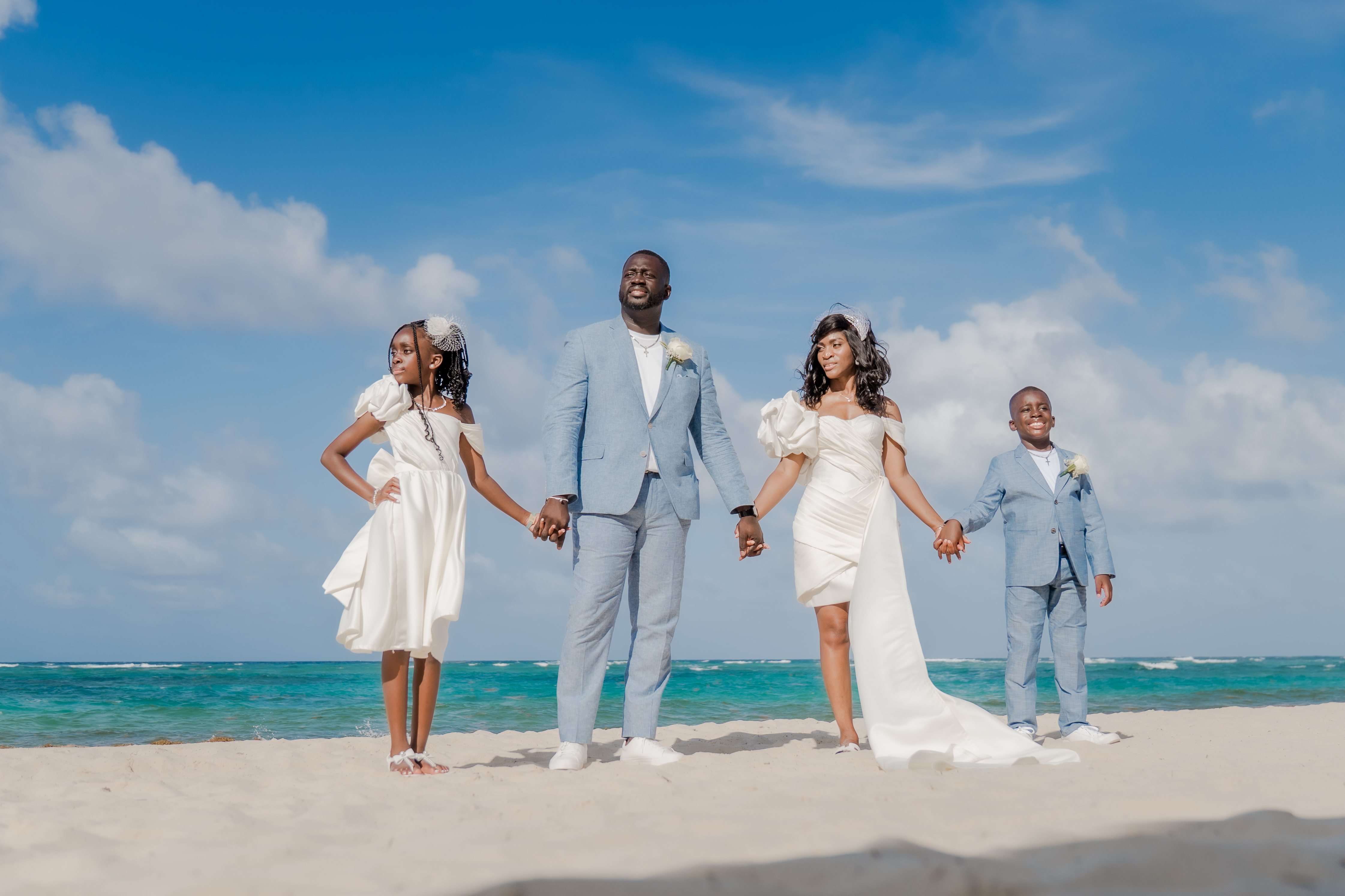 Four people in formal attire hold hands on a sandy beach at a destination wedding.