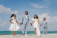 A family in formal attire holds hands on a beach at a destination wedding, ocean waves in the background.