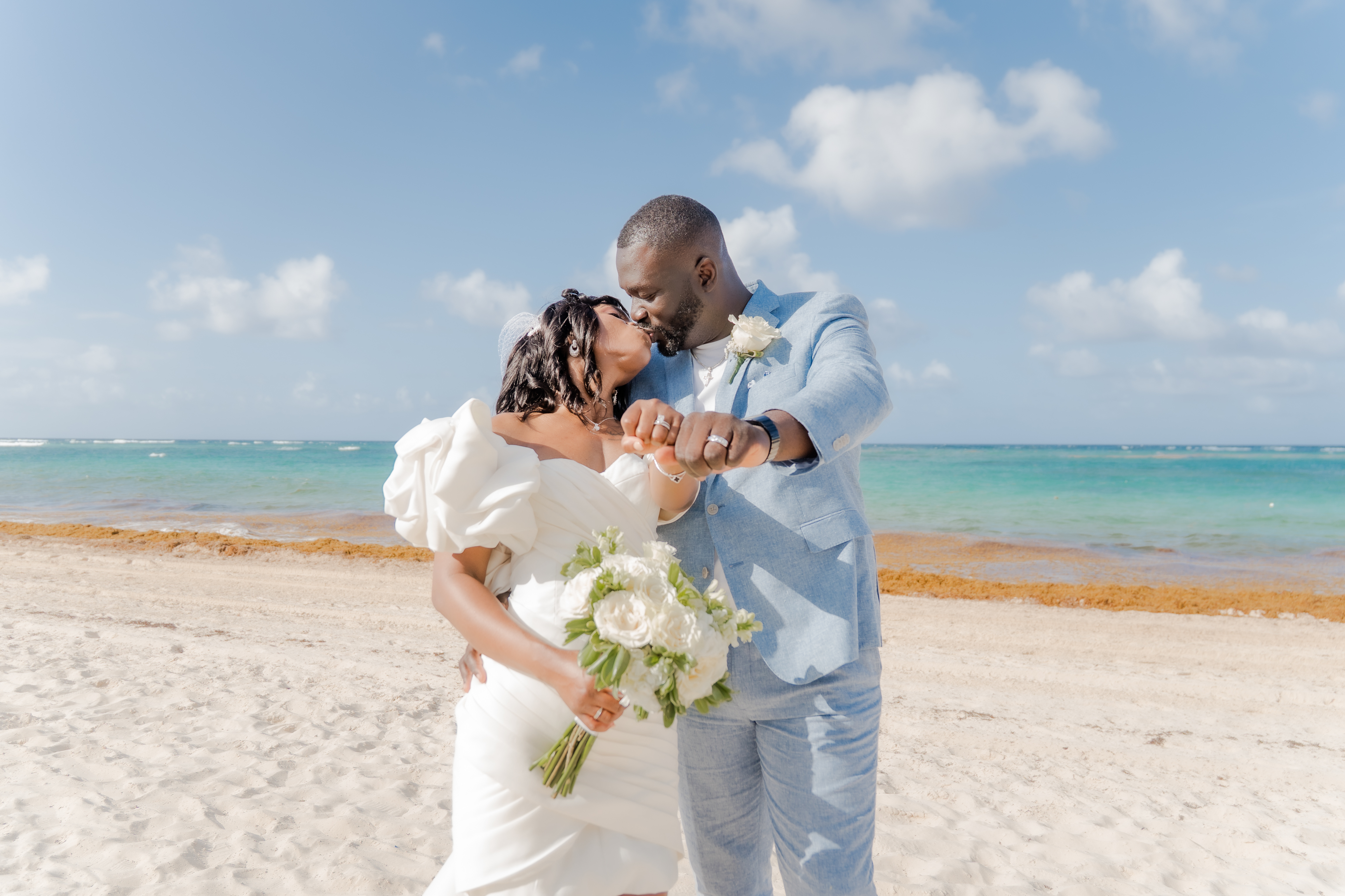Mallory and Kharey share a kiss on the sand during their destination wedding, bouquet and waves behind.