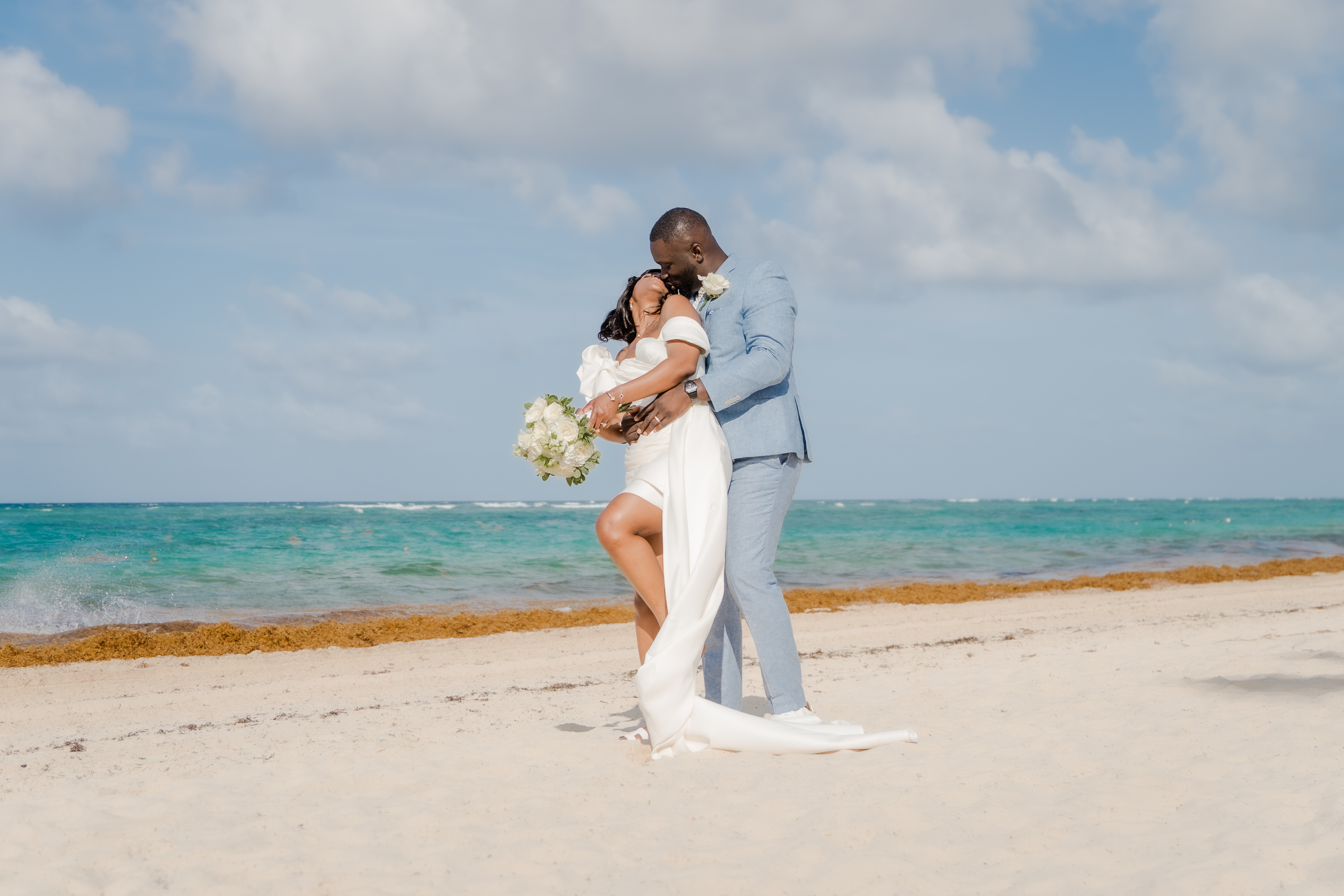 Mallory and Kharey (Copy) embrace on a sandy beach during their destination wedding by the ocean.