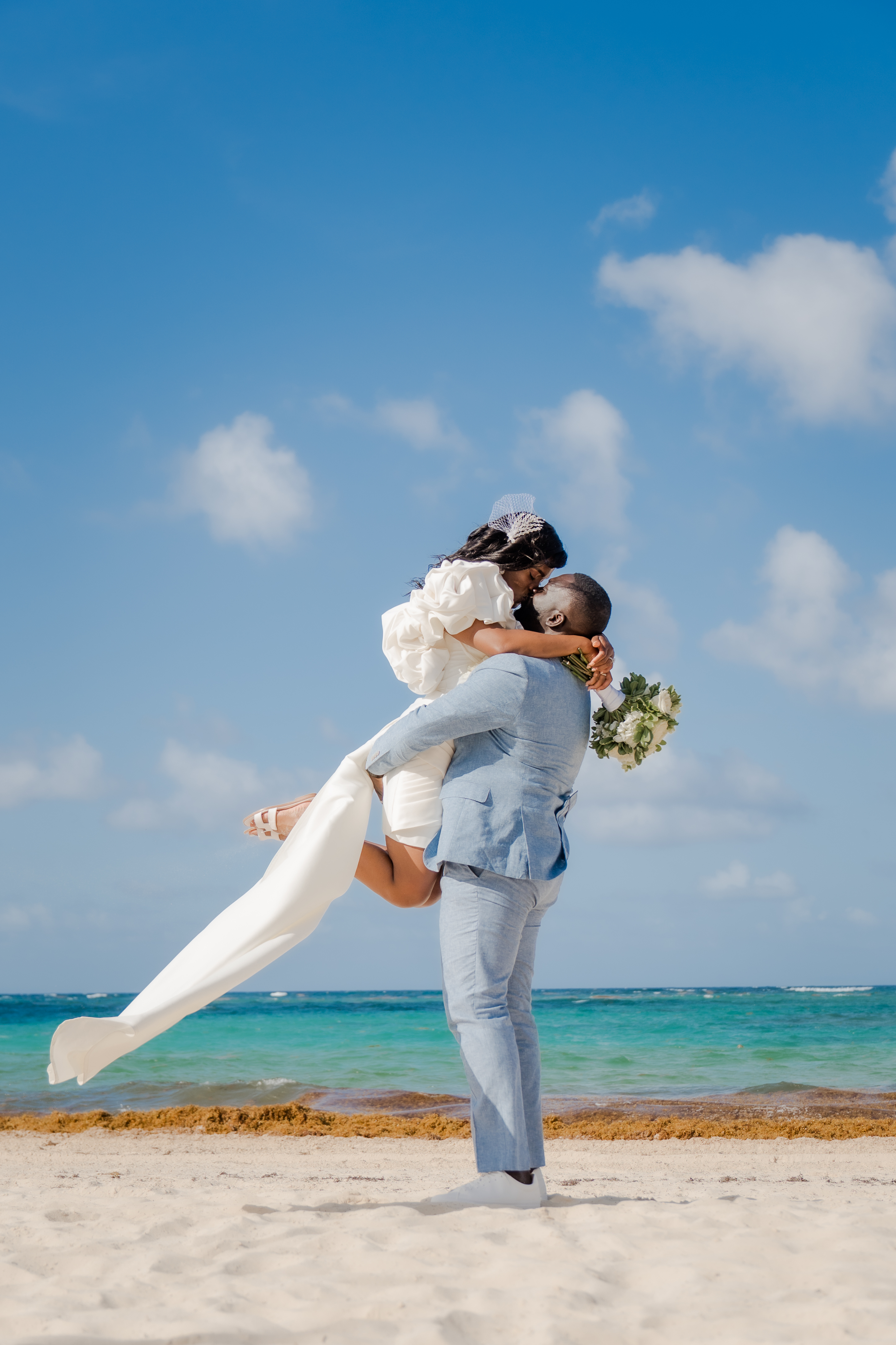 At their destination wedding, the groom lifts the bride on a sandy beach with ocean and blue sky behind.