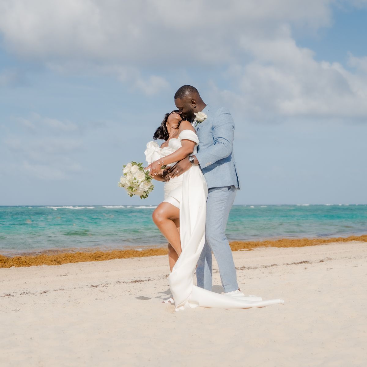 Mallory and Kharey embrace on a sandy beach at their destination wedding, ocean and sky behind them.
