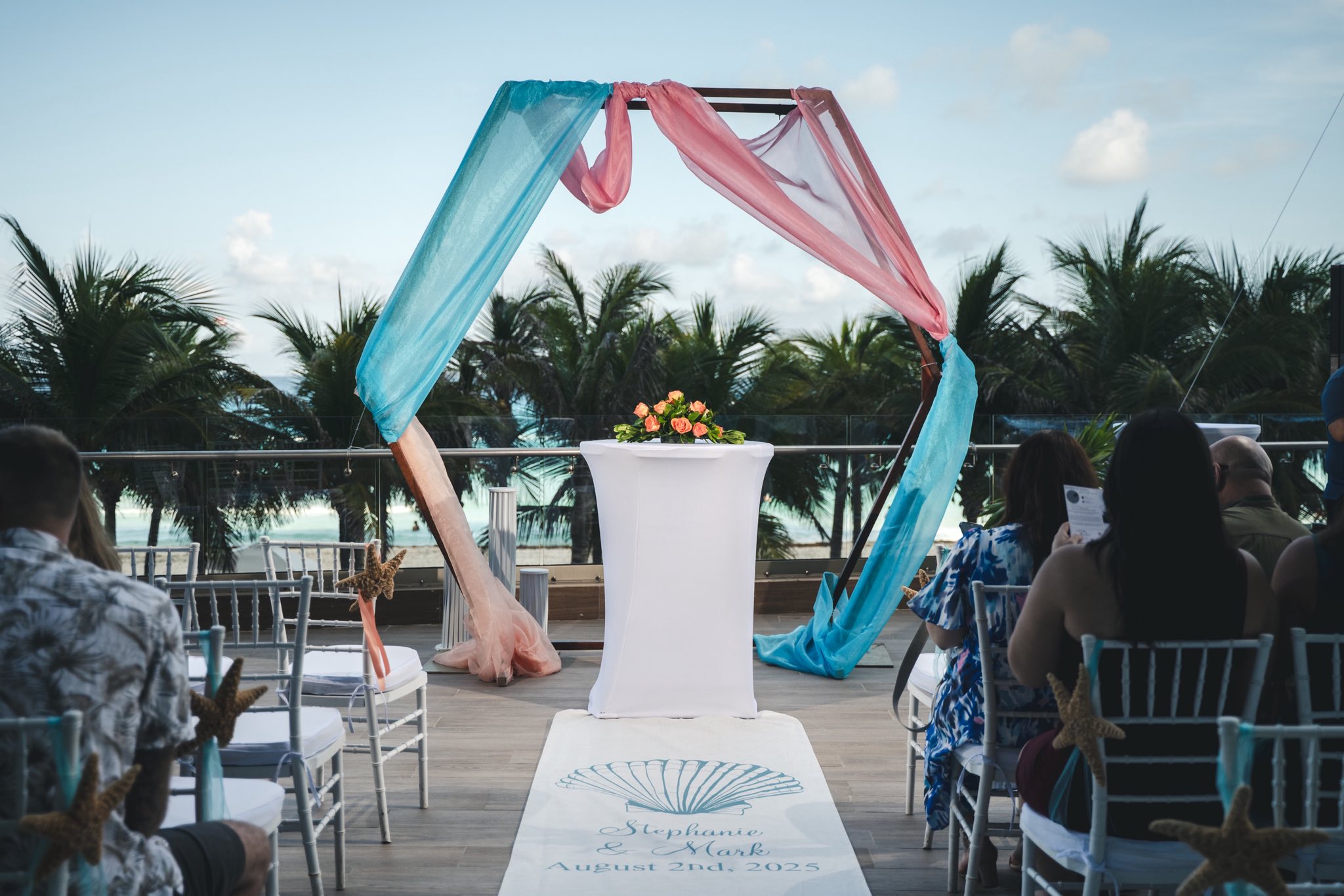 Mobolaji and Sylvia’s destination wedding outdoors, blue and pink floral arch, guests seated nearby.