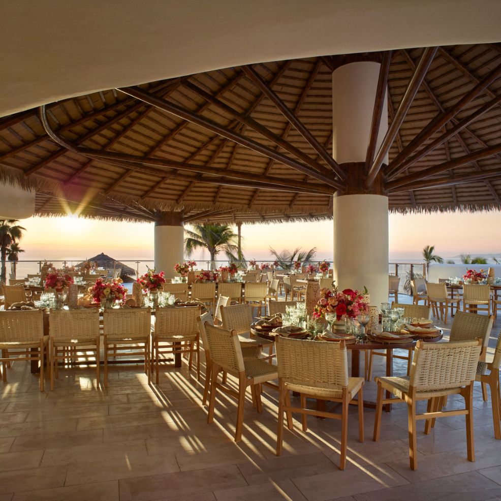 Open-air wedding restaurant with wooden tables, floral centerpieces, and sunset ocean view&mdash;perfect for a destination wedding.
