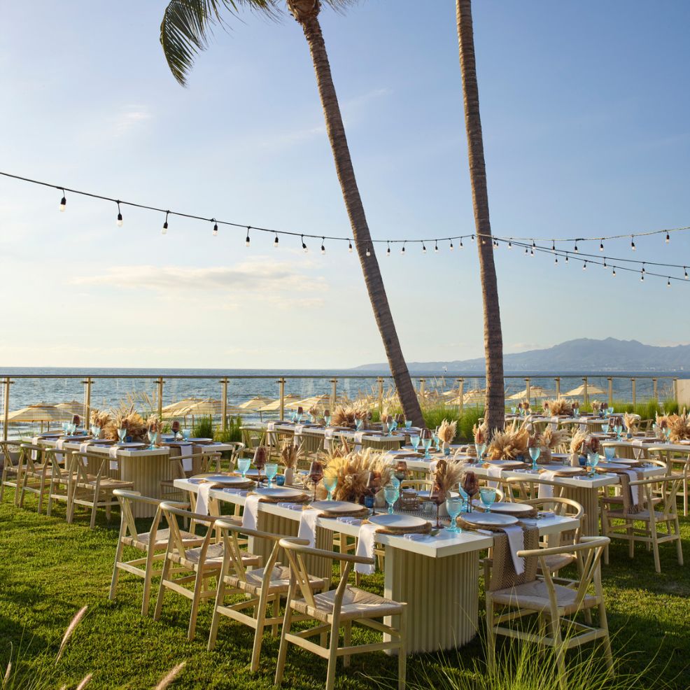 Destination wedding setup featuring elegantly decorated tables and chairs by the ocean under string lights.