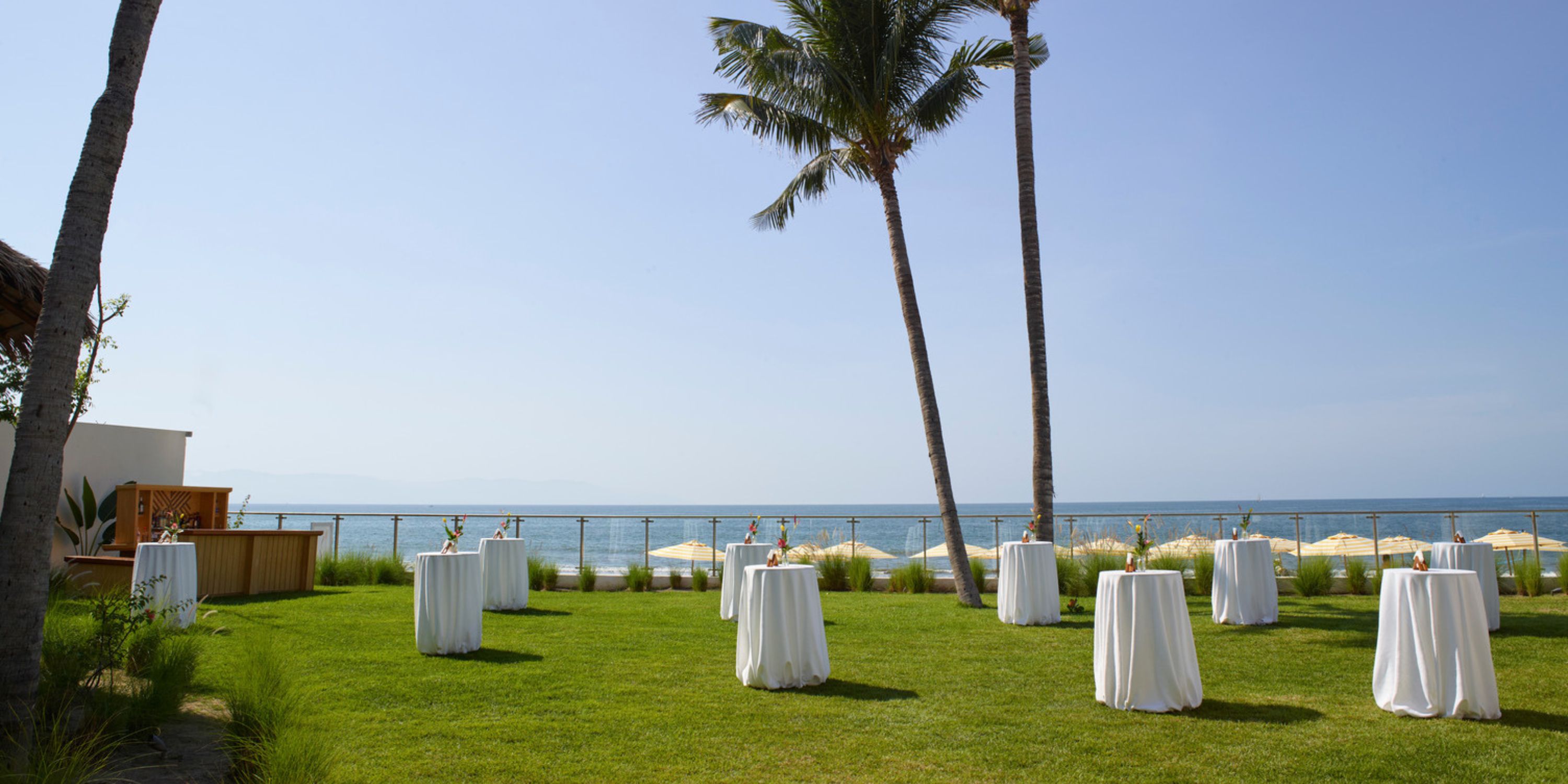 Cocktail tables with white cloths are arranged on a lawn by the ocean for a destination wedding.