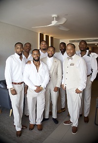 Eight men dressed in coordinated white and beige Jordan and Kievan formal attire are pictured indoors, preparing for a destination wedding.