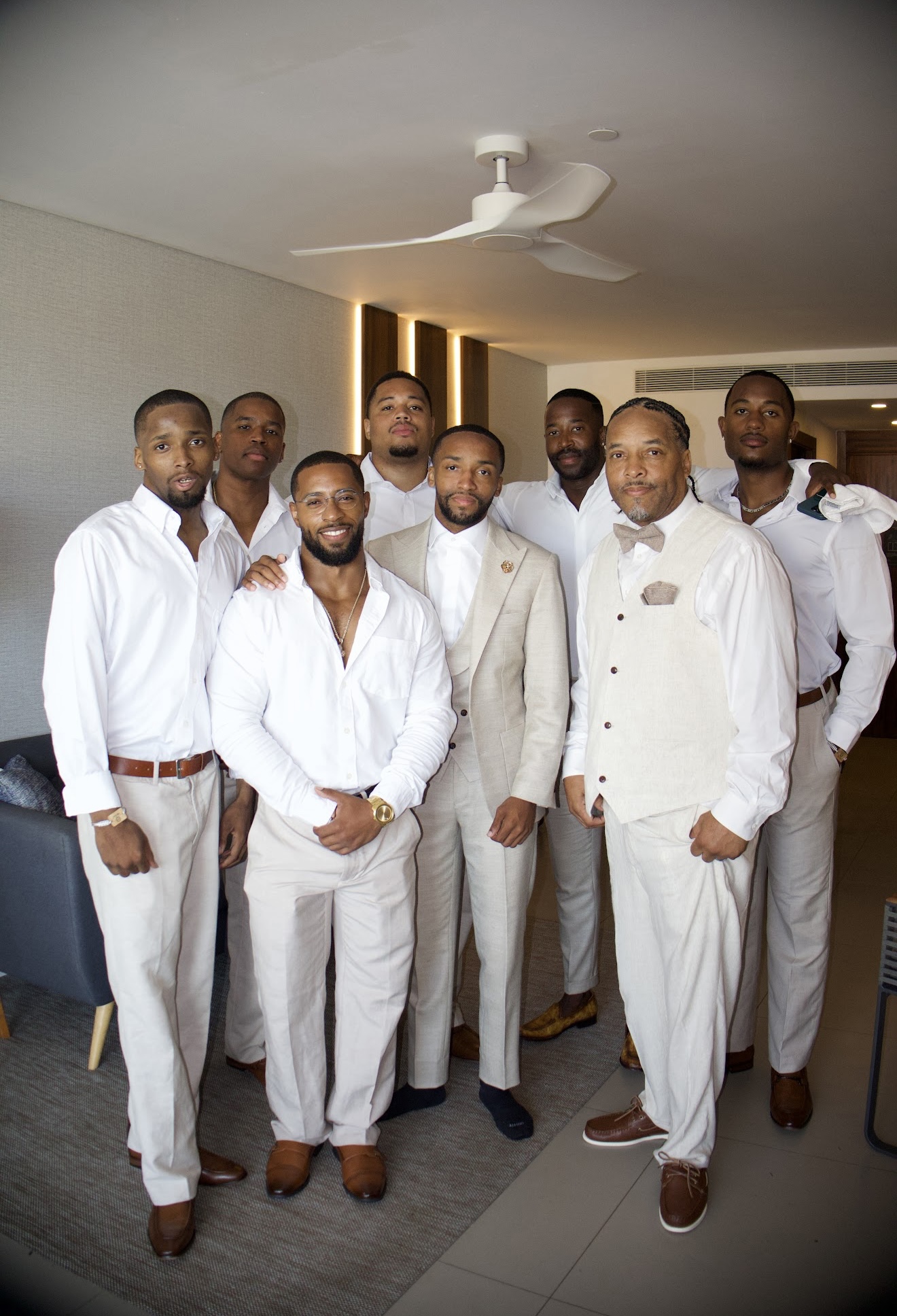 Eight men dressed in coordinated white and beige Jordan and Kievan formal attire are pictured indoors, preparing for a destination wedding.