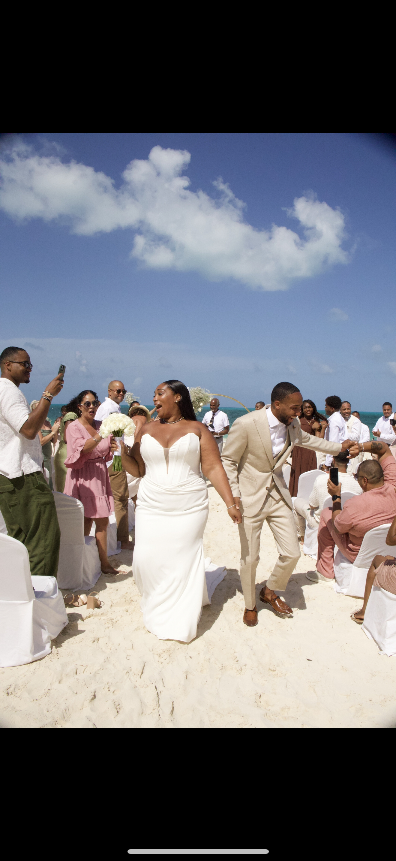 Jordan and Kievan walk hand in hand down a sandy beach aisle at their destination wedding, while guests eagerly snap photos.