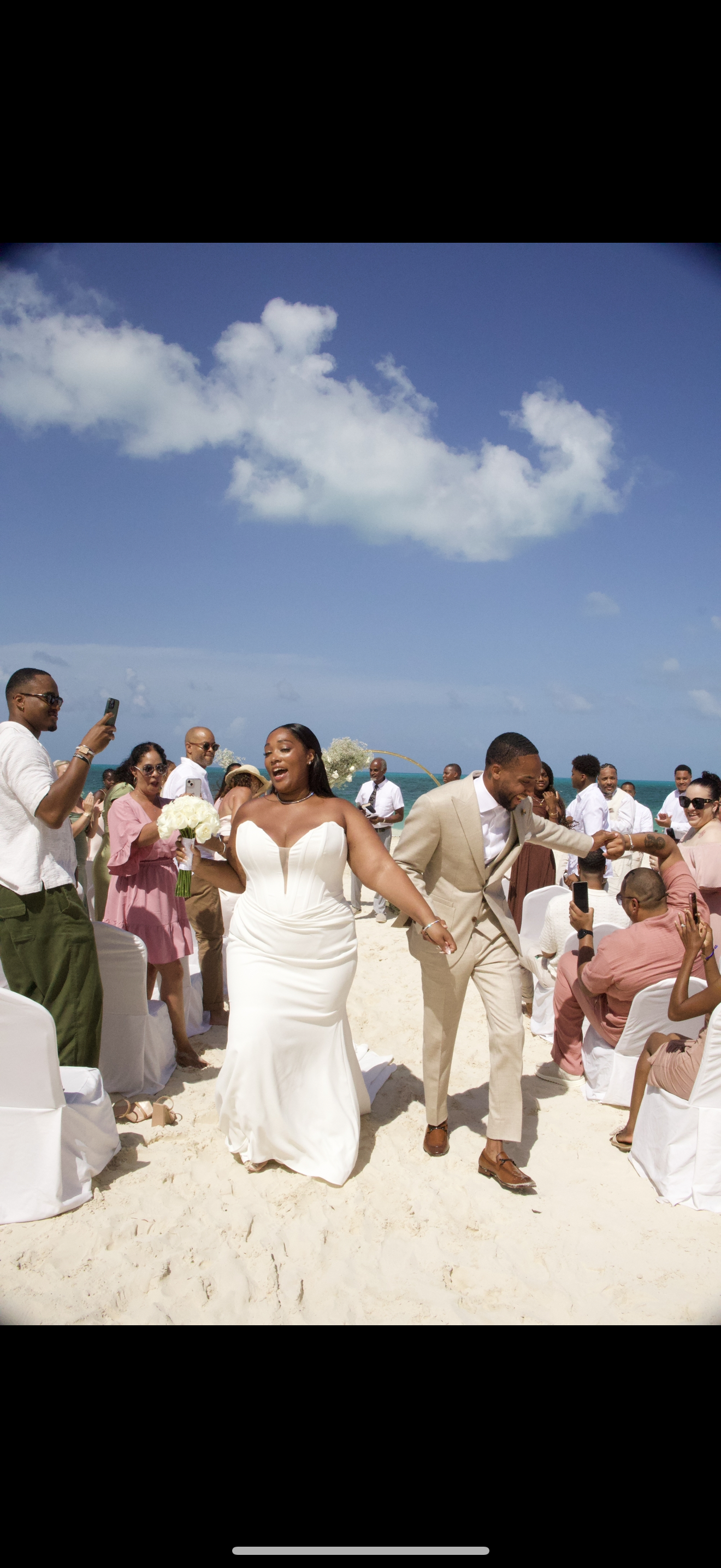 Jordan and Kievan walk down the aisle at their destination wedding on a sandy beach, surrounded by their guests.