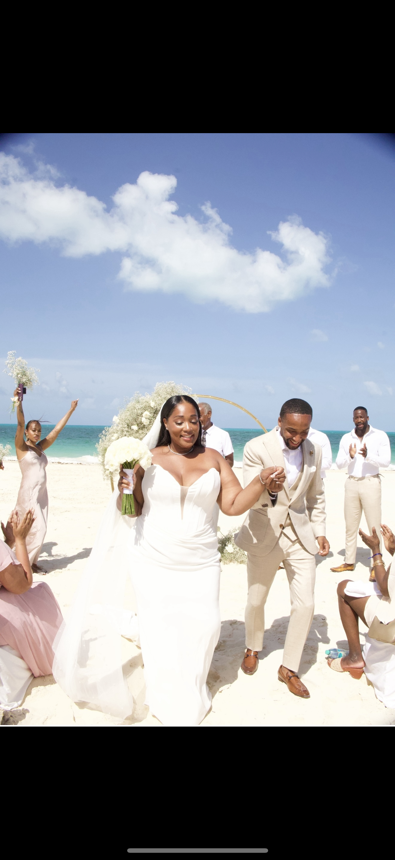 Jordan and Kievan stroll along the beach, celebrating their destination wedding with their bridal party under clear blue skies.