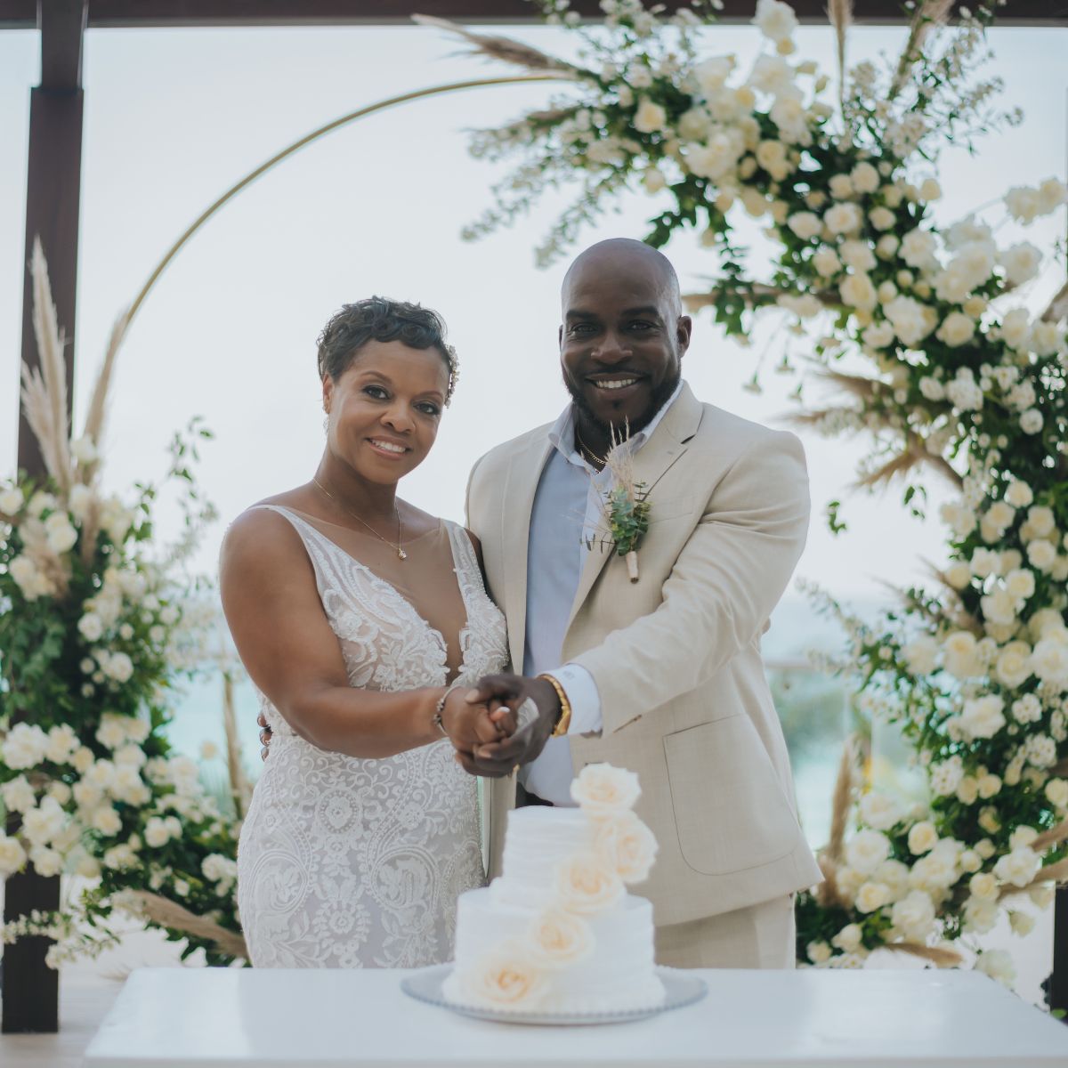 A couple in formal wear cuts their wedding cake in front of a floral arch at a destination wedding.